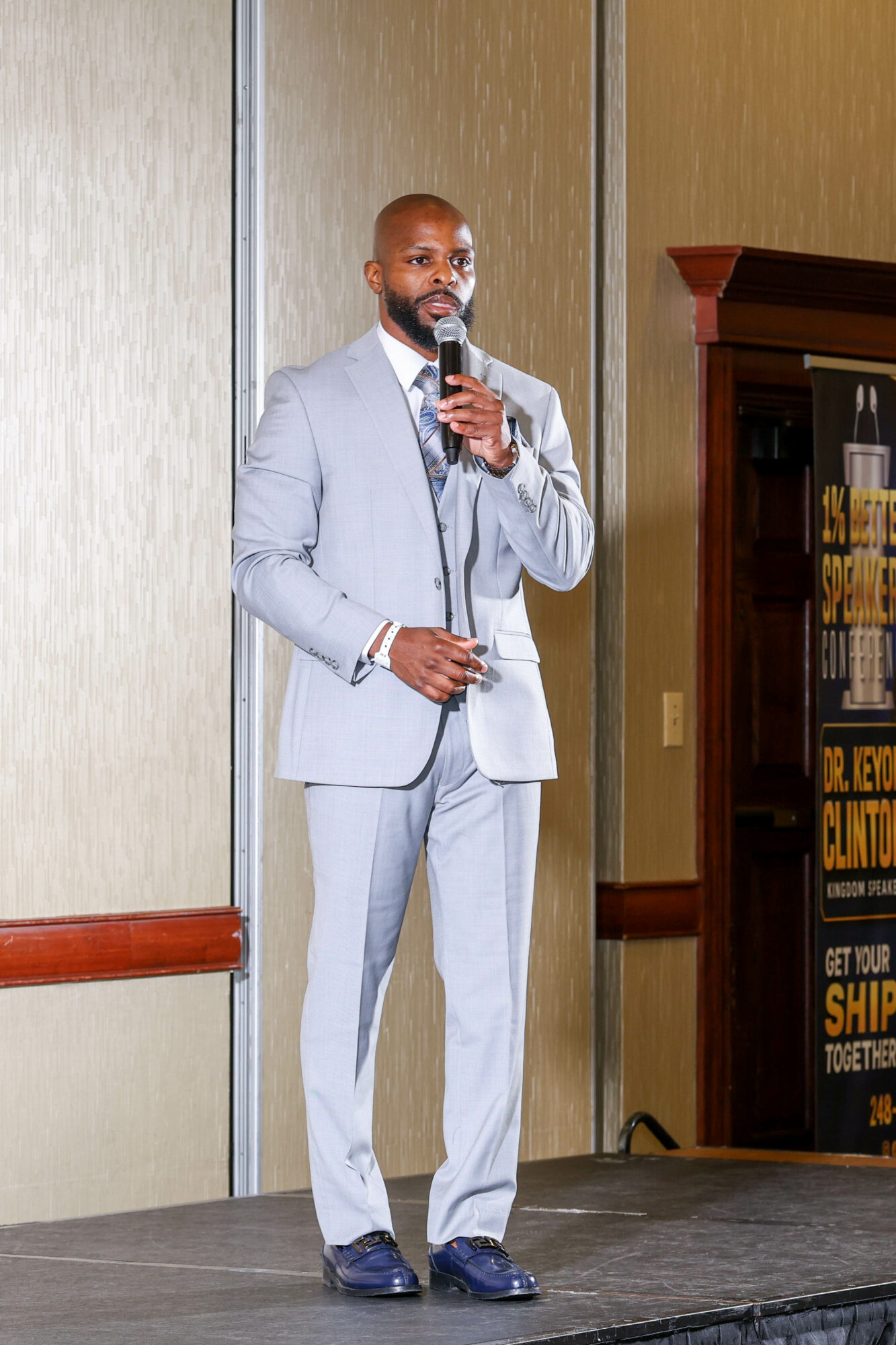 Man in light gray suit speaking into a microphone on stage, with a wooden cabinet and beige wall behind him.