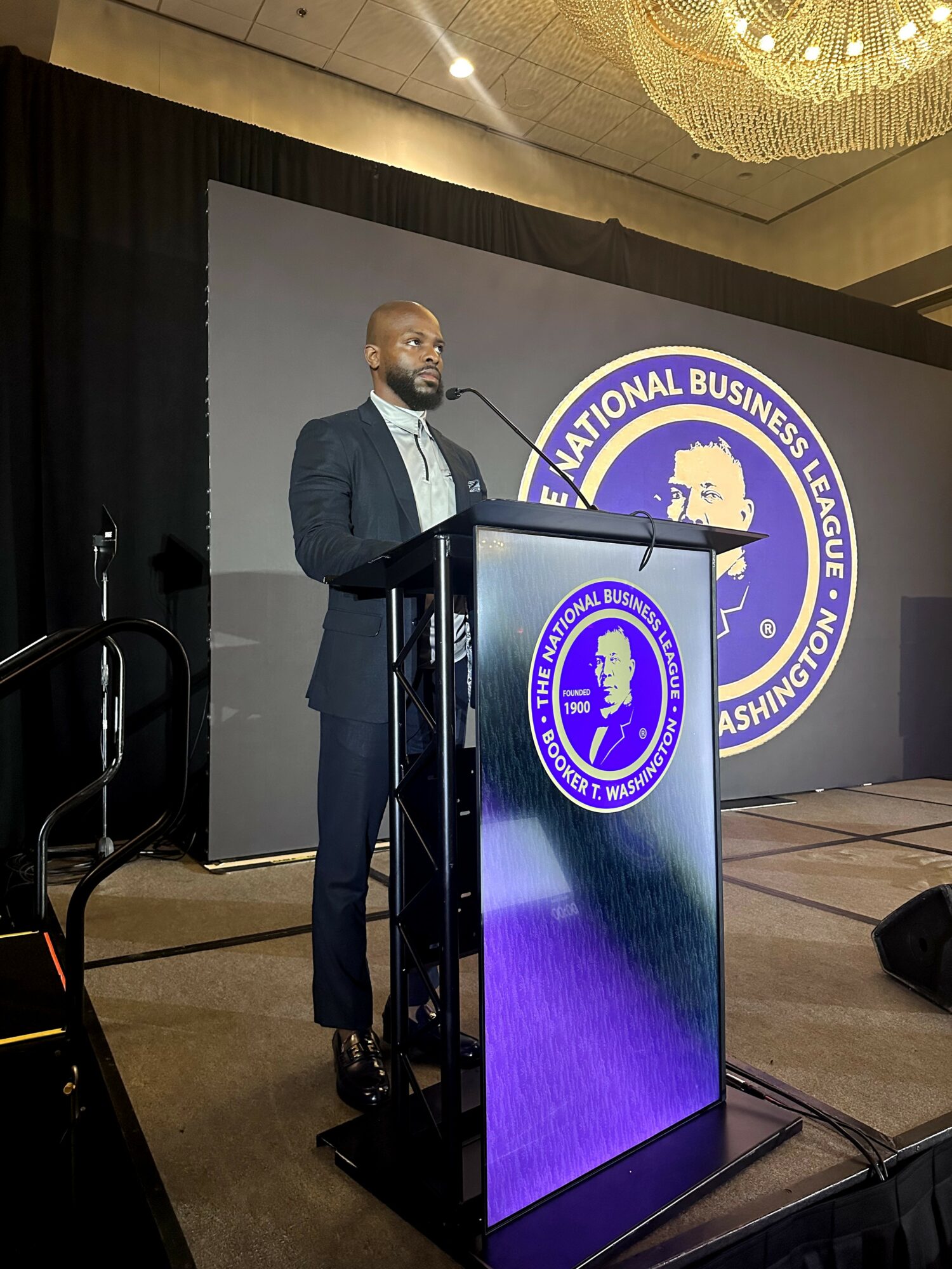 Man in suit speaking at a podium during a conference, with a large logo behind him reading 'National Business League' and 'Washington, D.C.'