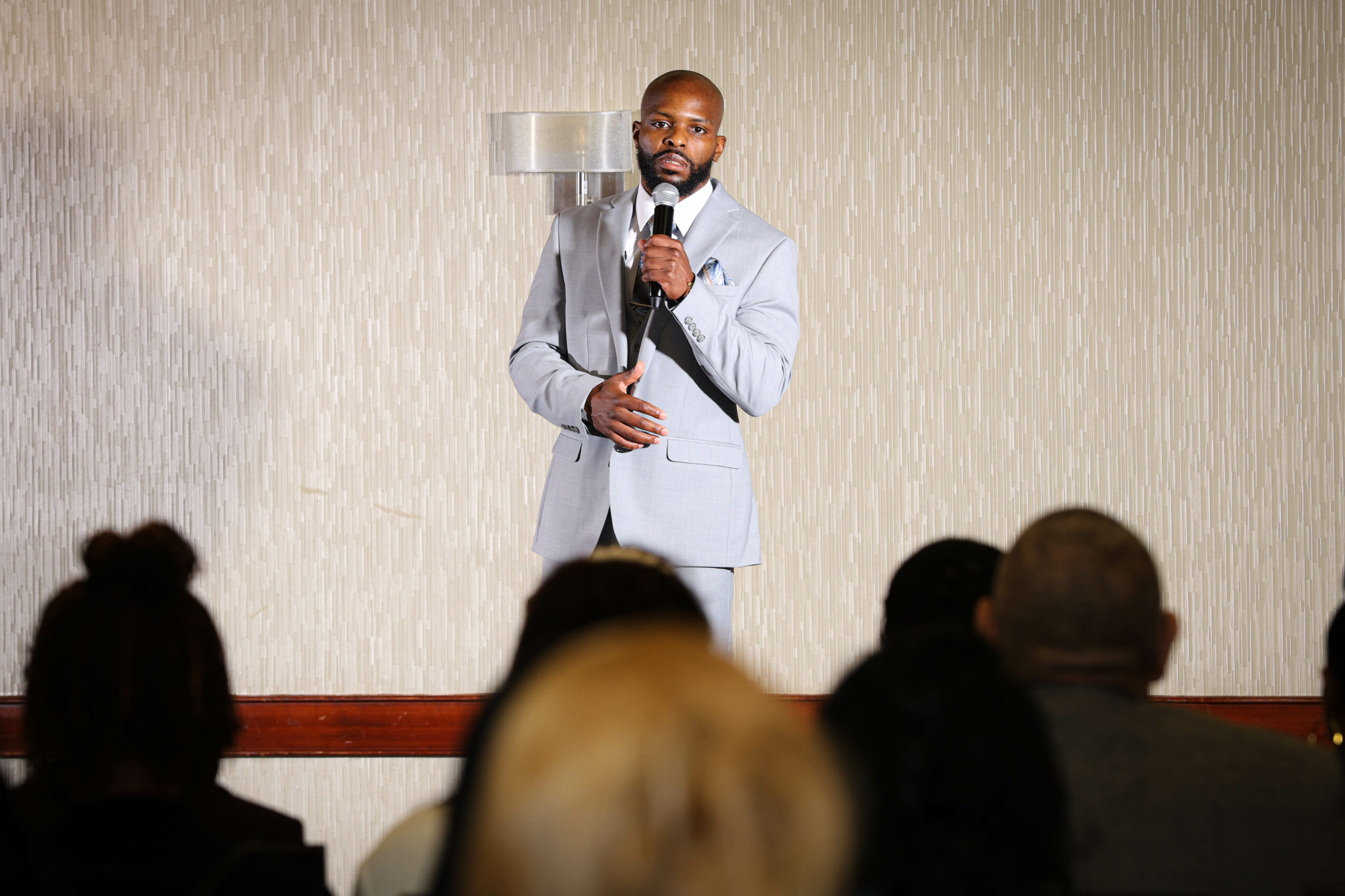 Man in light gray suit speaking into microphone in front of audience, beige wall background.