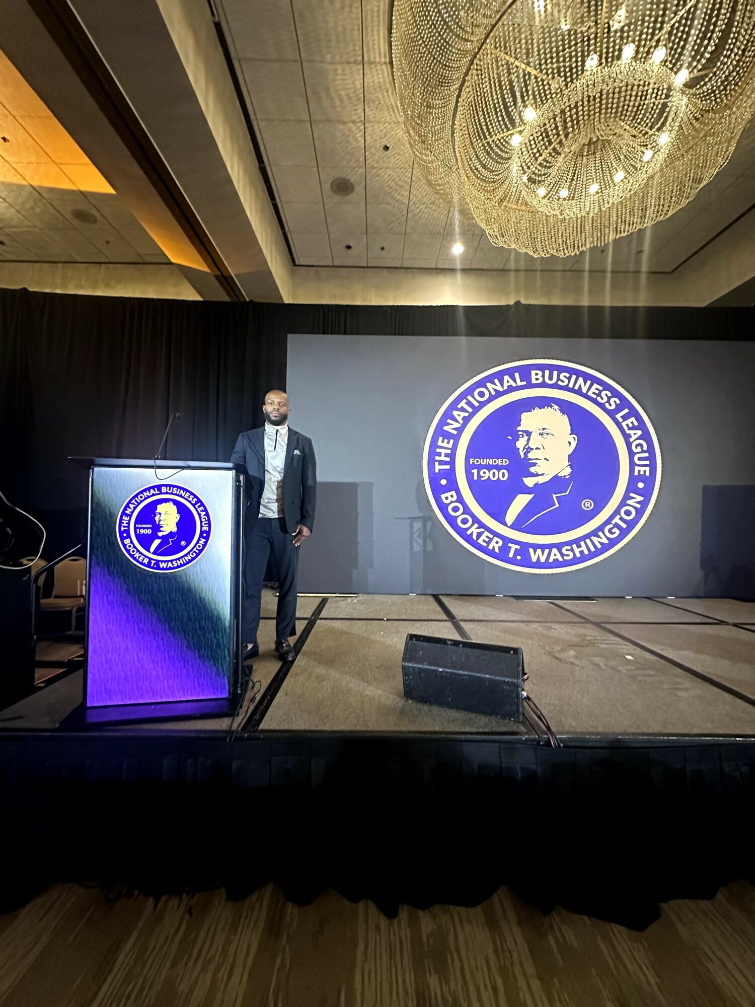 Person standing on stage with a large logo of Booker T. Washington on the screen behind, in a conference room with a chandelier.