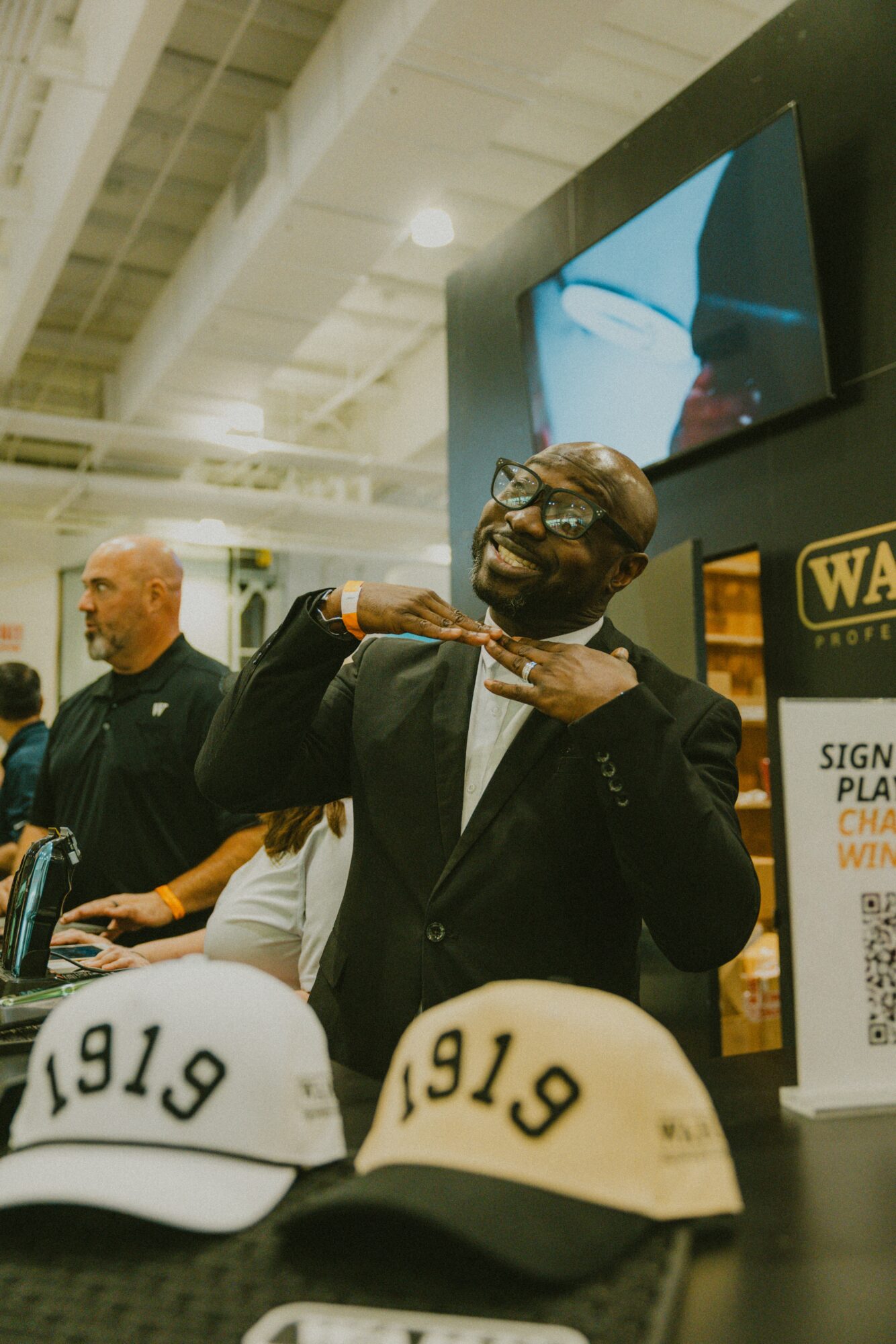 Person in suit and glasses posing with hand on neck at event, hats with '1919' in front, indoor setting.