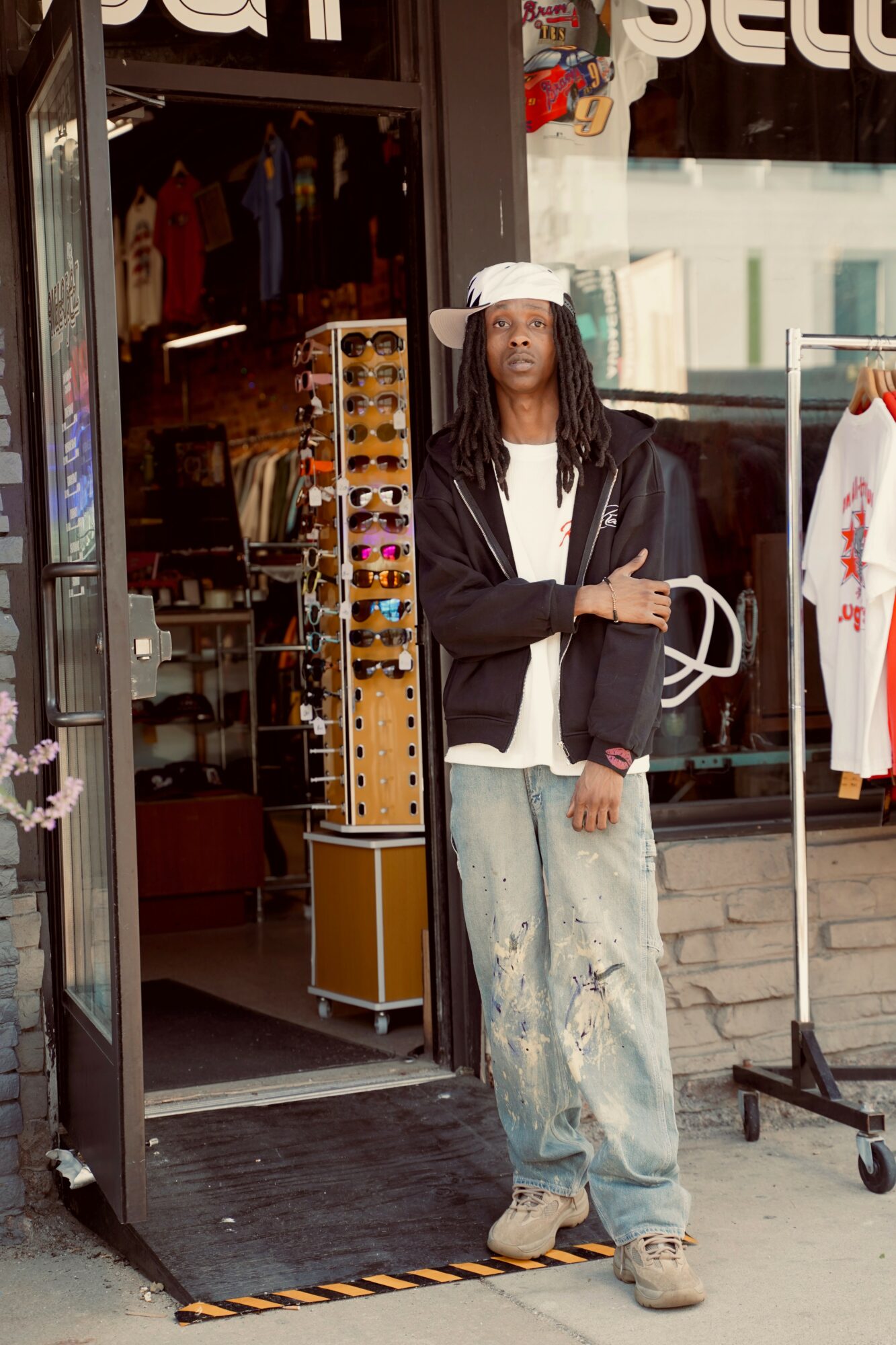 Person with long hair, white cap, black jacket, and ripped jeans standing outside a store entrance.