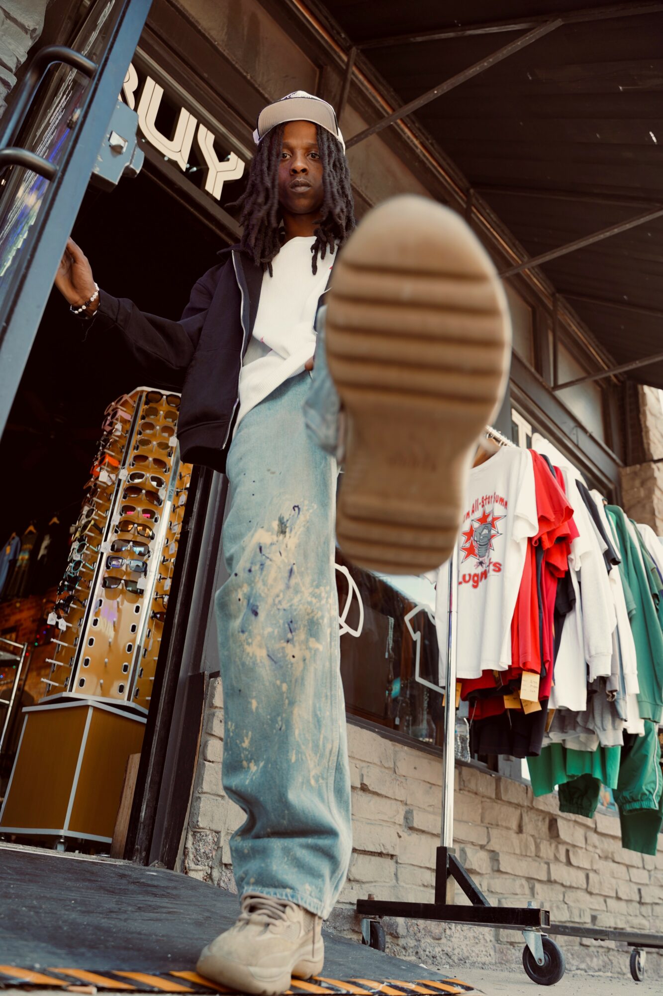 Person standing outside a store, foot raised towards camera, clothing displayed behind, wearing a cap and casual outfit.