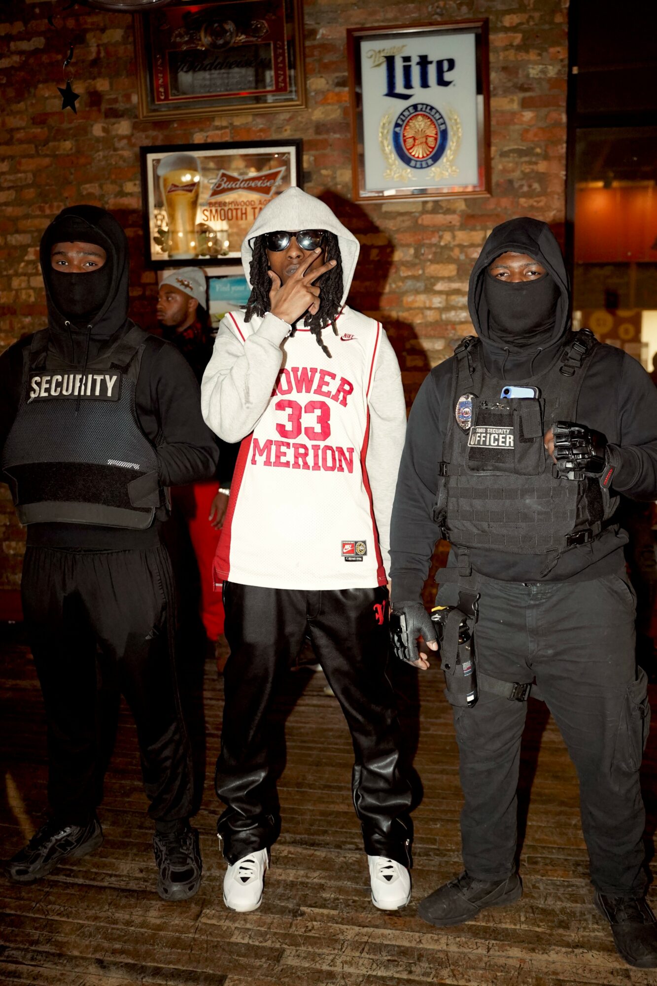 Three people in dark clothing and masks stand in front of a brick wall with framed pictures and a Miller Lite sign.