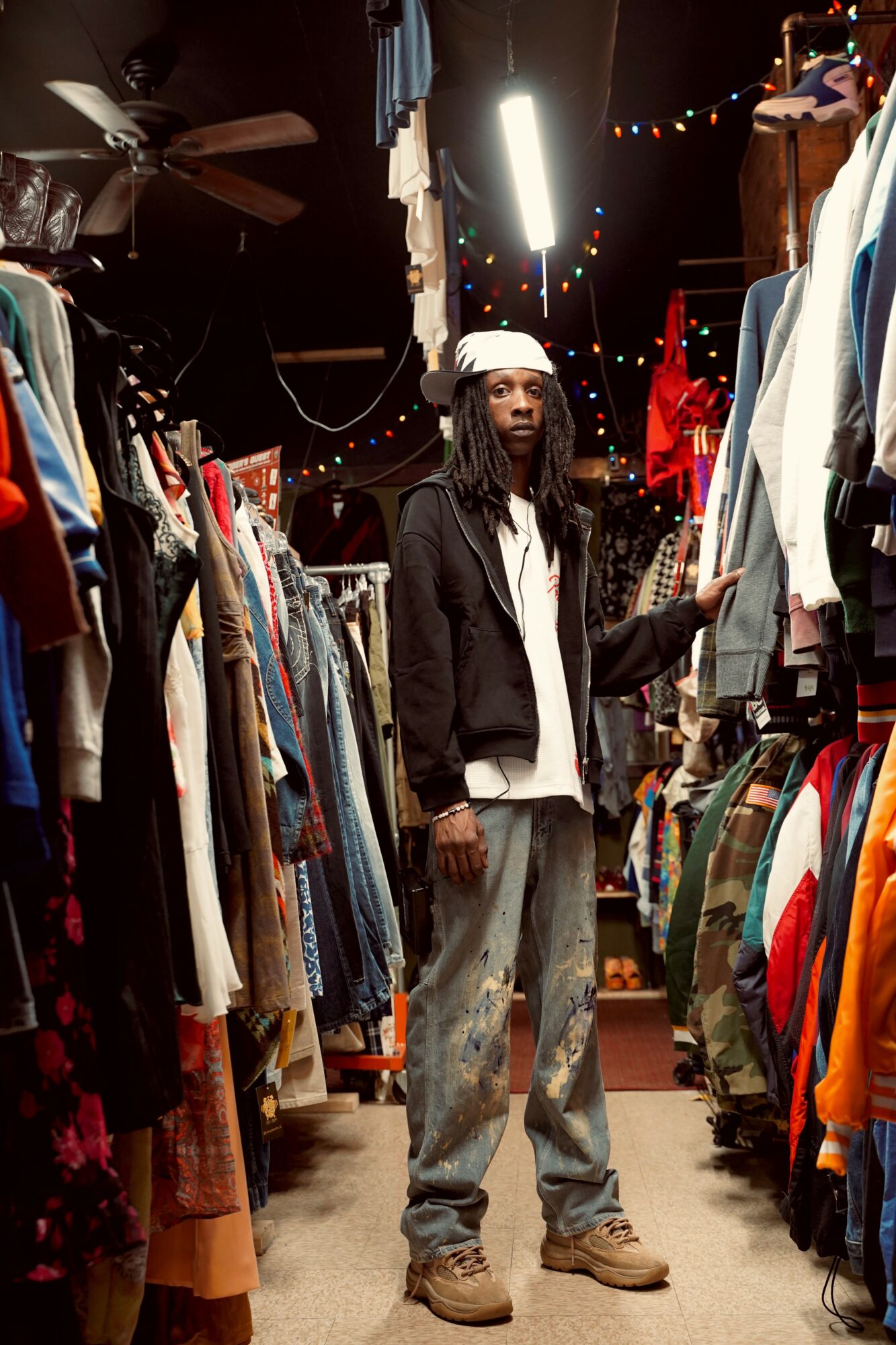 Person with dreadlocks wearing a white hat, black jacket, and jeans shopping among racks of clothes in a store.