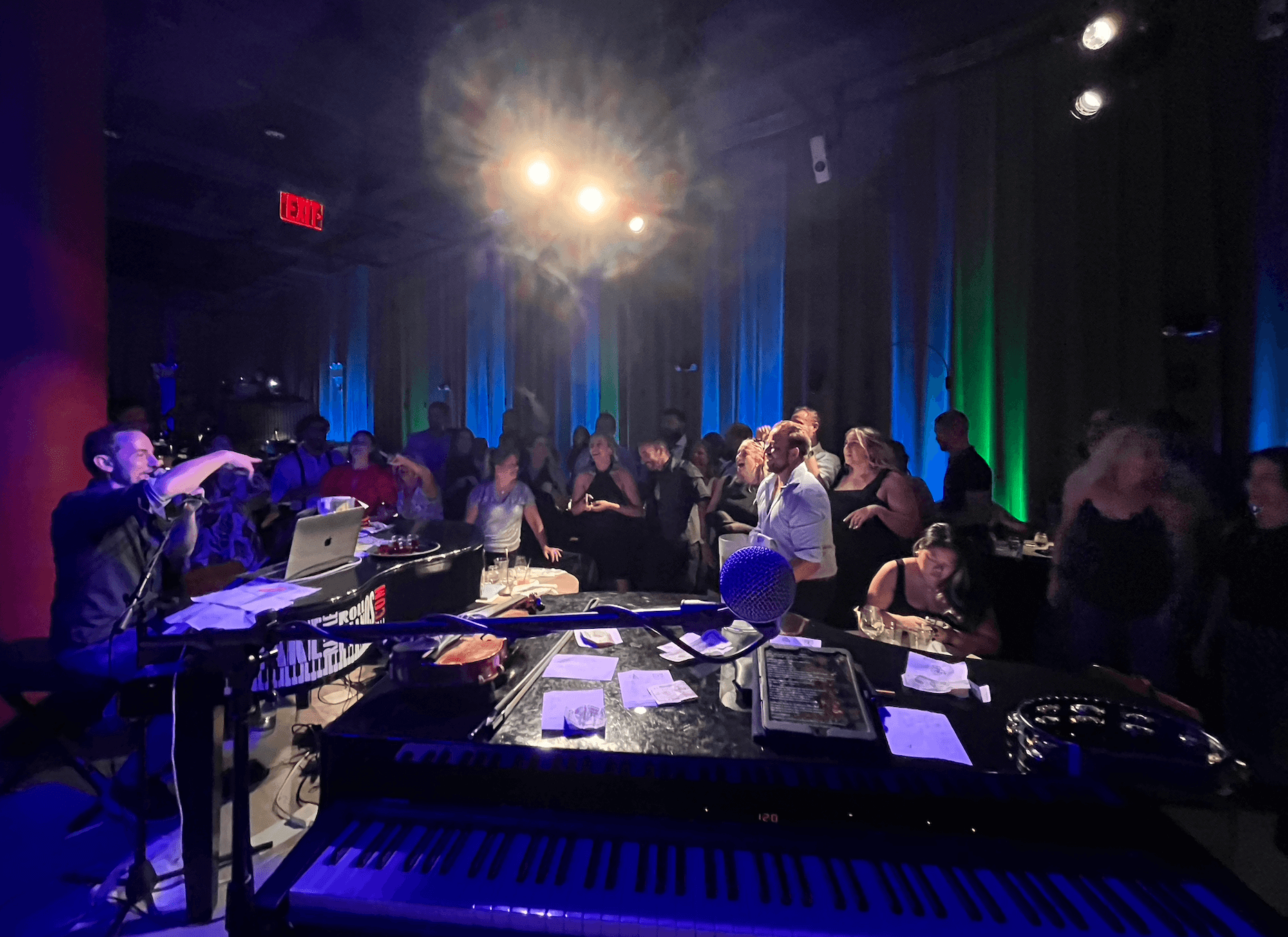 People gathered in a dimly lit room with colorful lighting, a musician playing keyboard, and a bright light overhead.