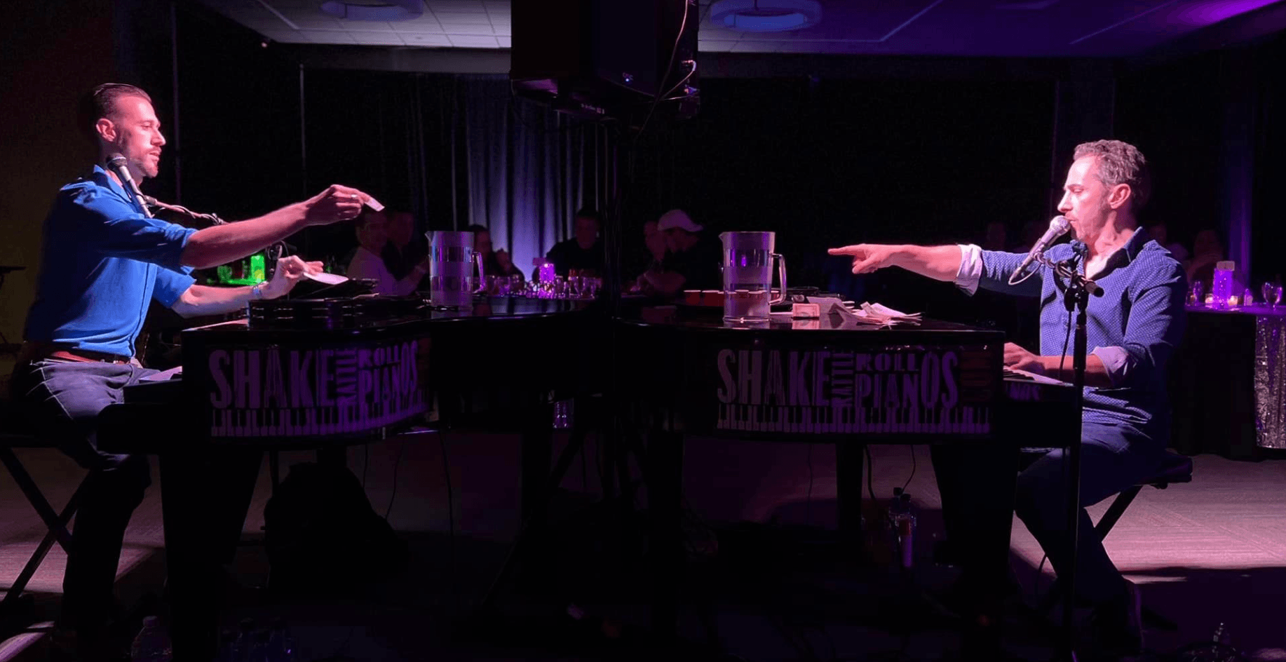 Two people sit at a table on a stage, reaching towards each other with extended arms, in a dark setting with purple lighting.