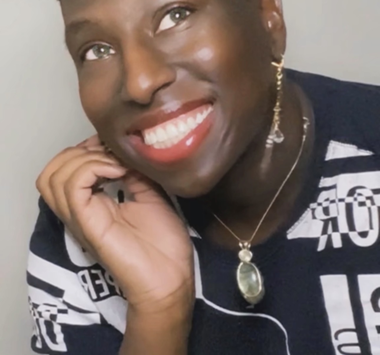 Close-up of a smiling person with short hair, wearing earrings, a necklace, and a patterned top, resting their chin on their hand.