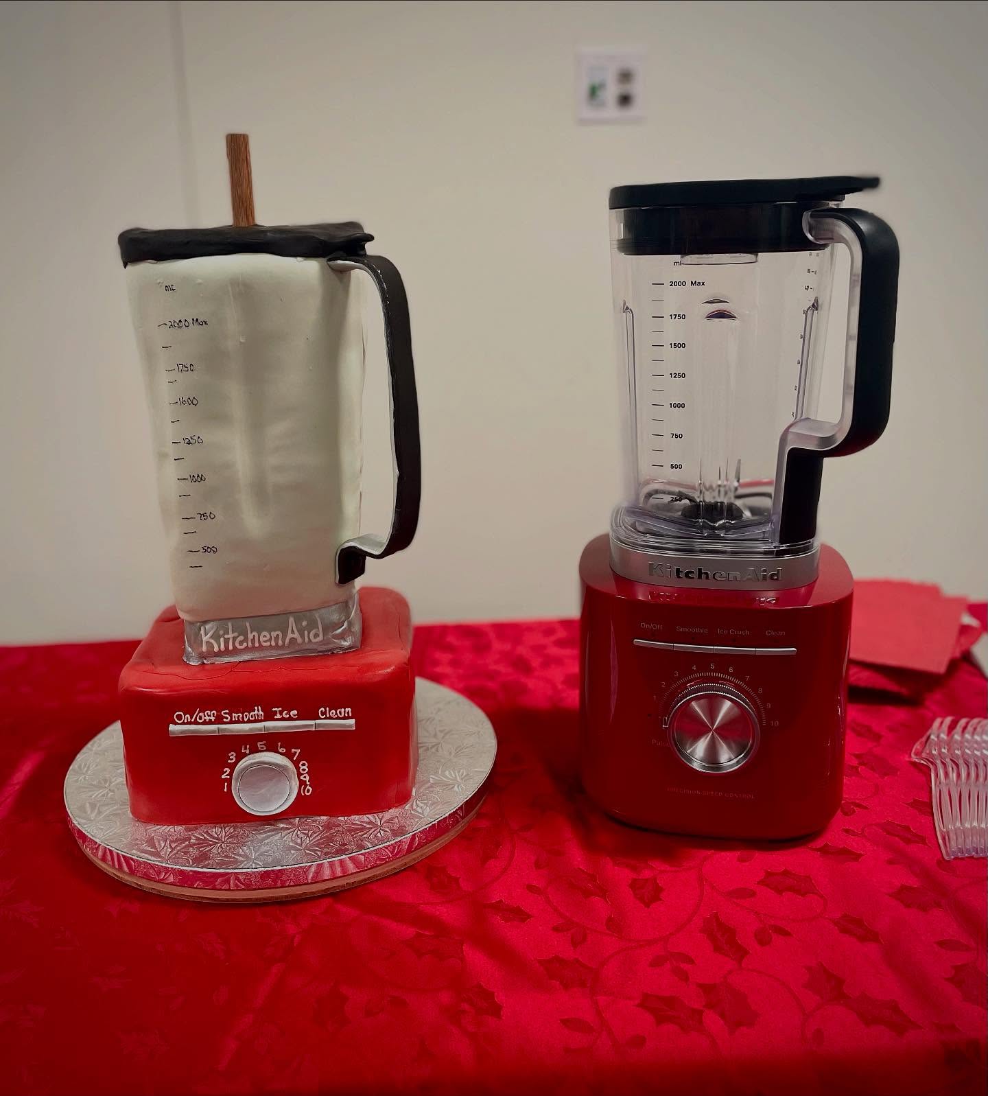 Two blenders on a red tablecloth, one with a measuring jug and the other with a glass container, in a room with a white wall.