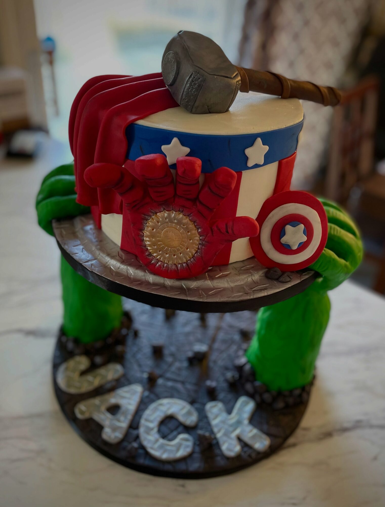 Cake decorated with a hammer, stars, and shield, on a wooden stand with green legs, with the name 'JACK' at the base.