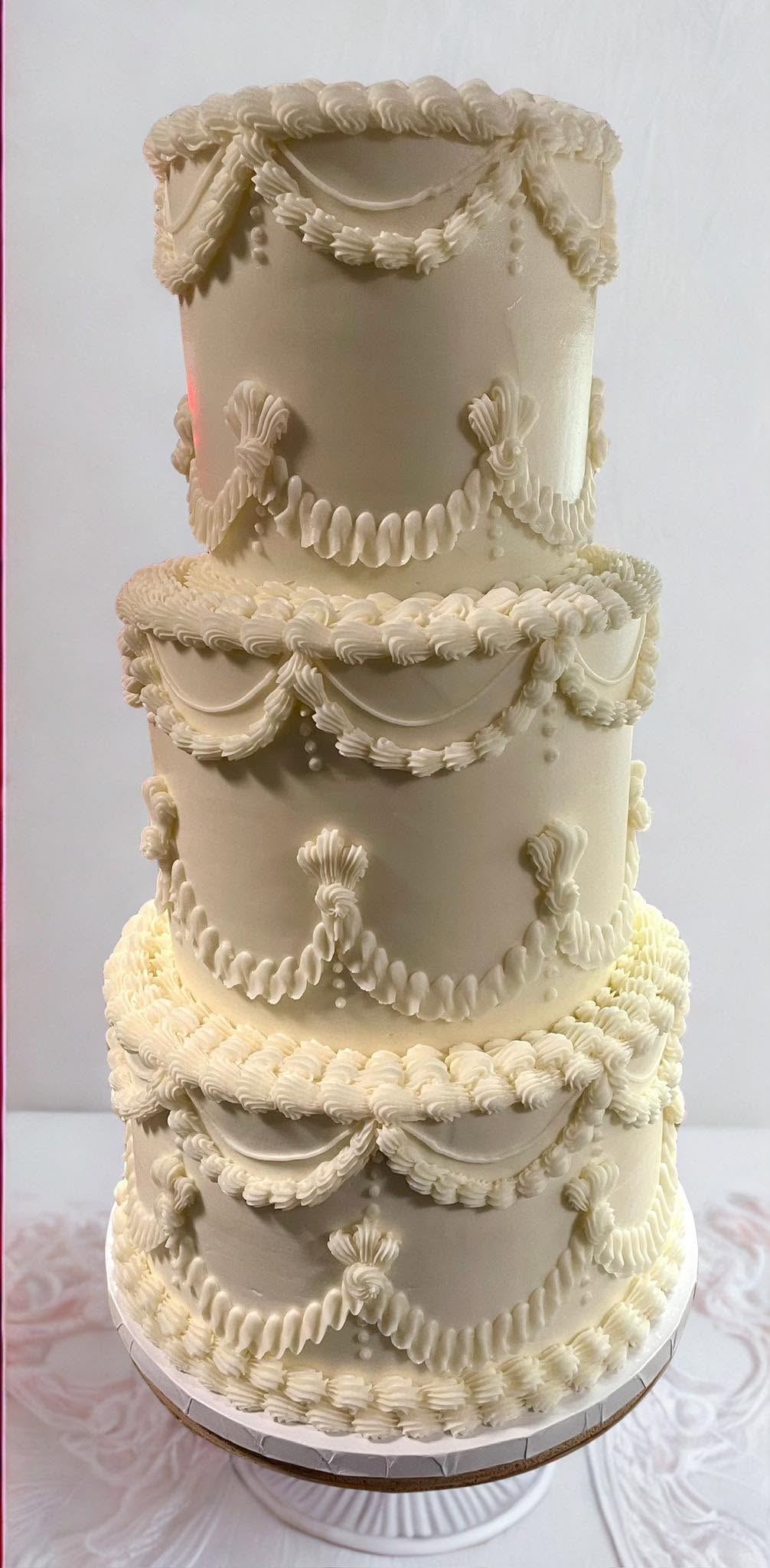 Four-tiered cake decorated with cream-colored garlands and swags, on a round base, against a plain background.