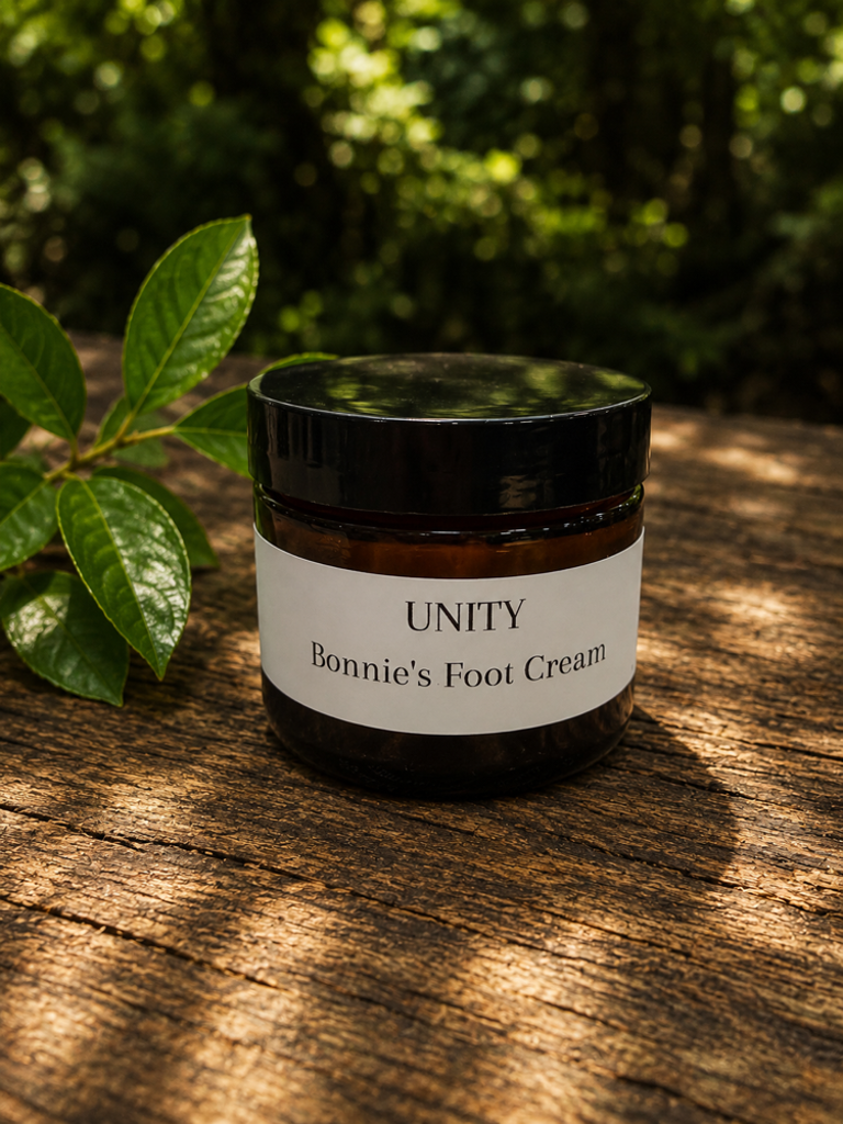 Brown jar of Bonnie's Foot Cream labeled 'UNITY' on a wooden surface with green leaves nearby, outdoors with trees in background.