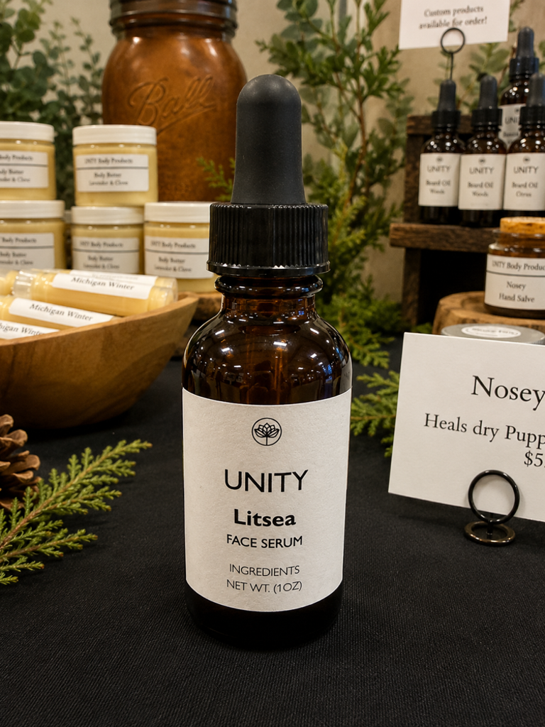 Small amber glass dropper bottle labeled 'UNITY' and 'Litsea Face Serum' on a black surface with herbal products in background.