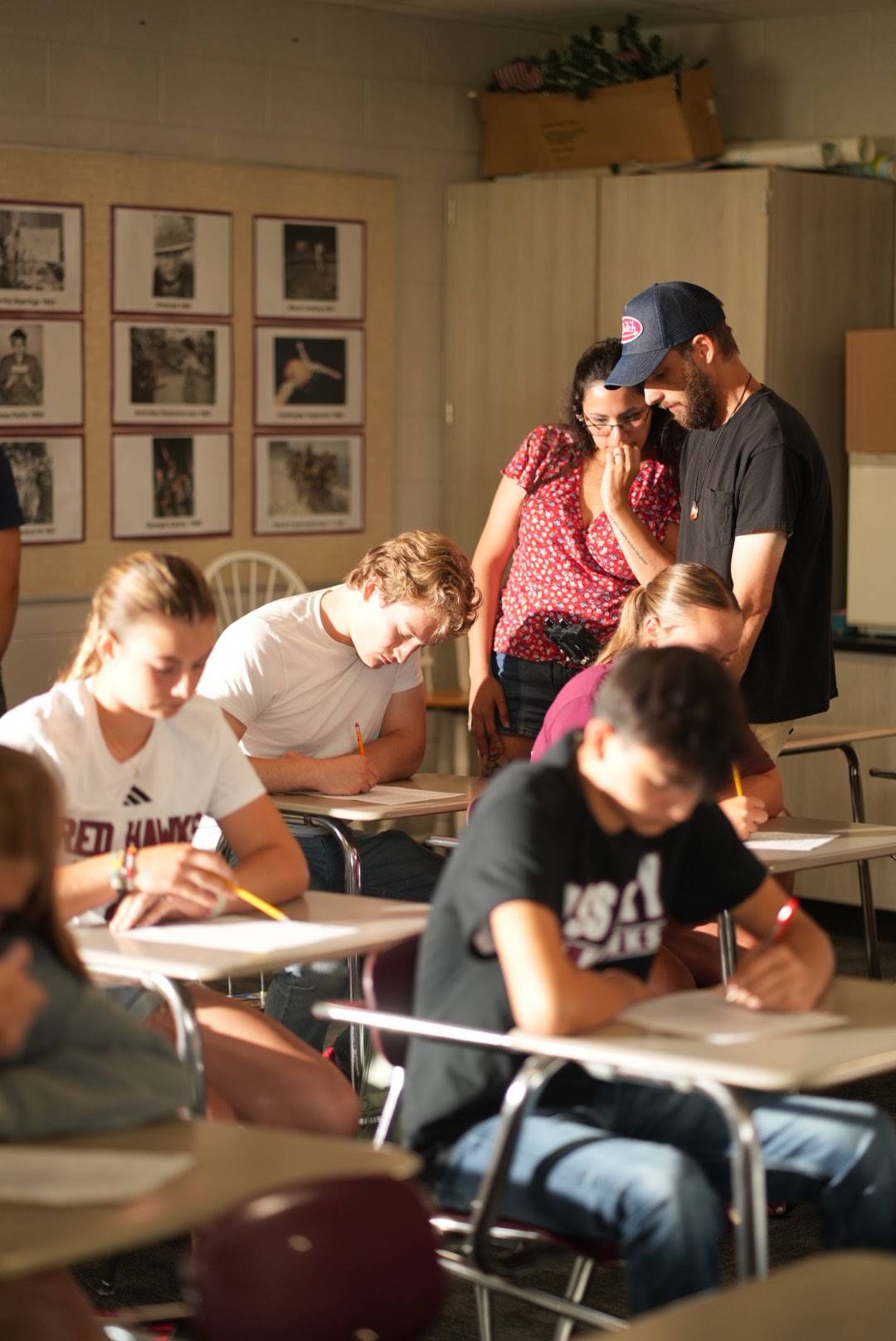 Students sit at desks in a classroom, writing, with two adults standing and talking in the background, wall with framed pictures.