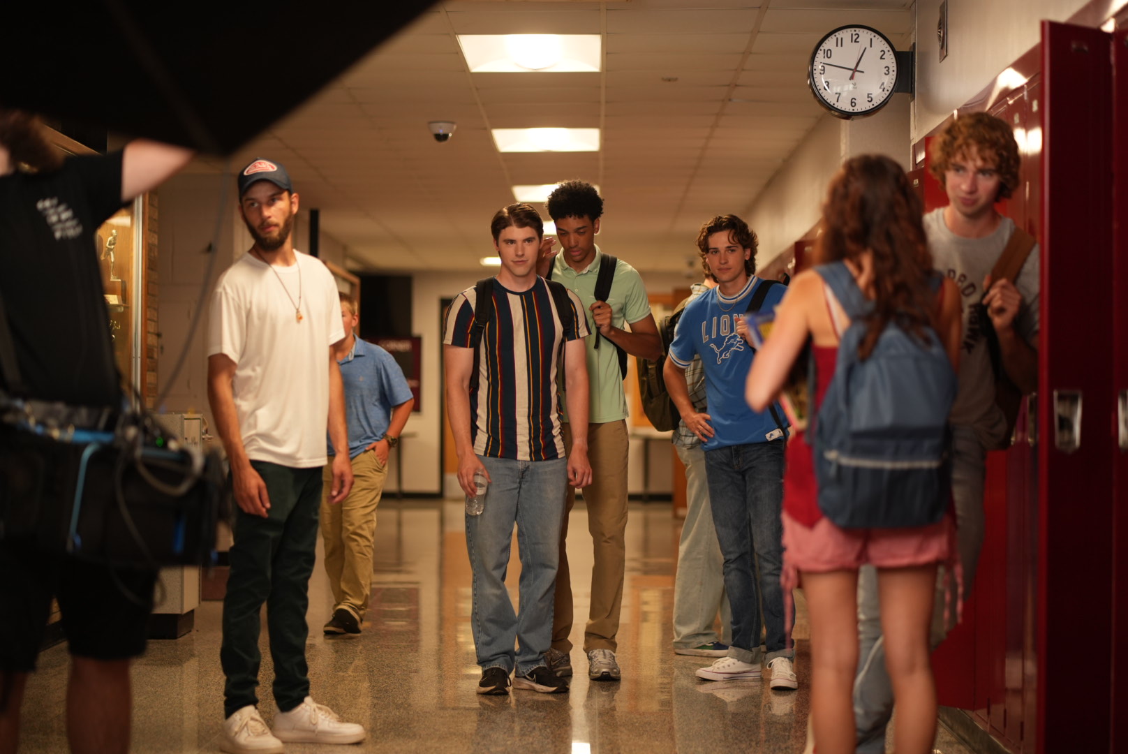 Group of students in a school hallway near lockers, some talking, some looking at a person with camera equipment, clock on wall.