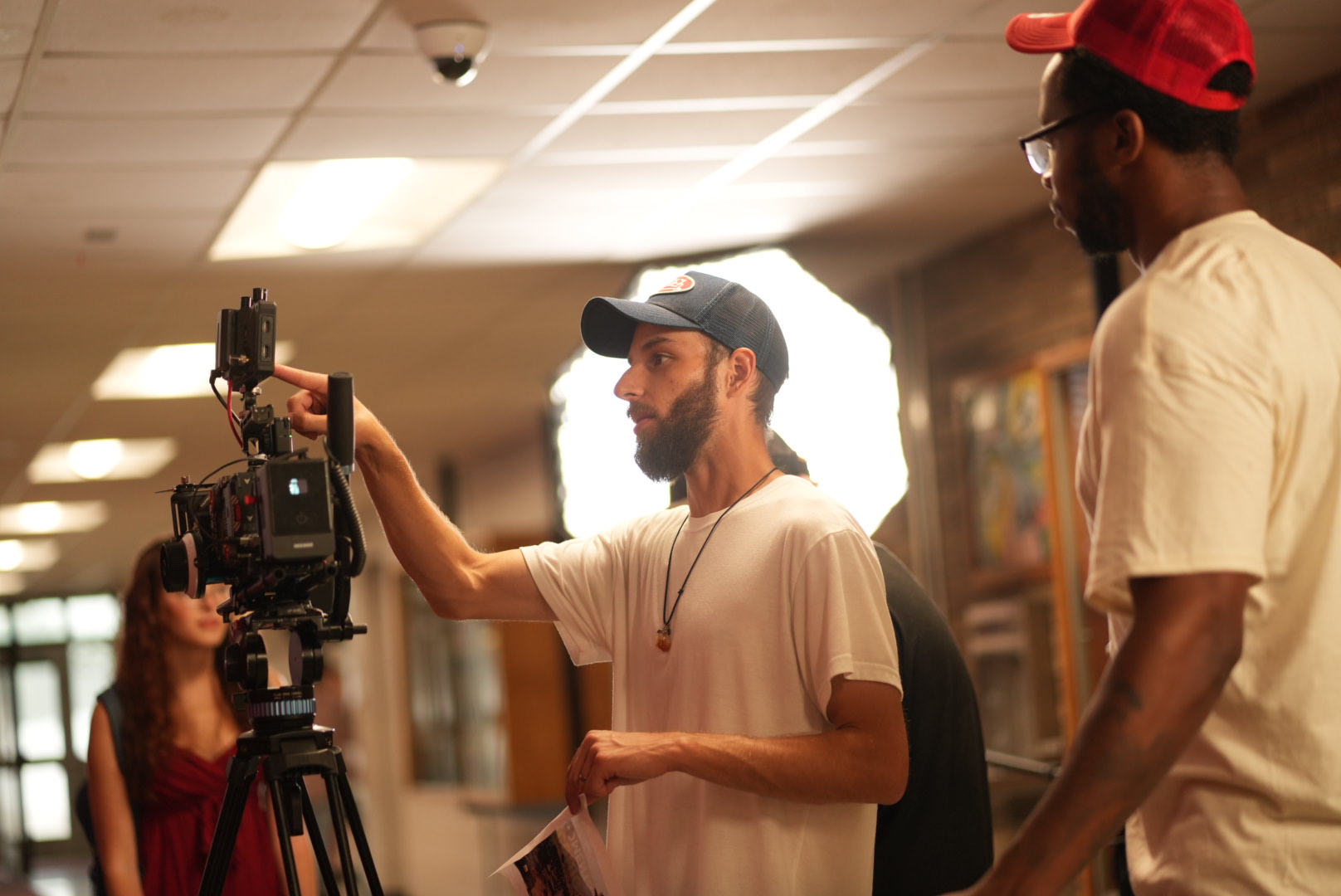 Two men and a woman on a film set, with one man adjusting camera equipment, in a room with warm lighting.