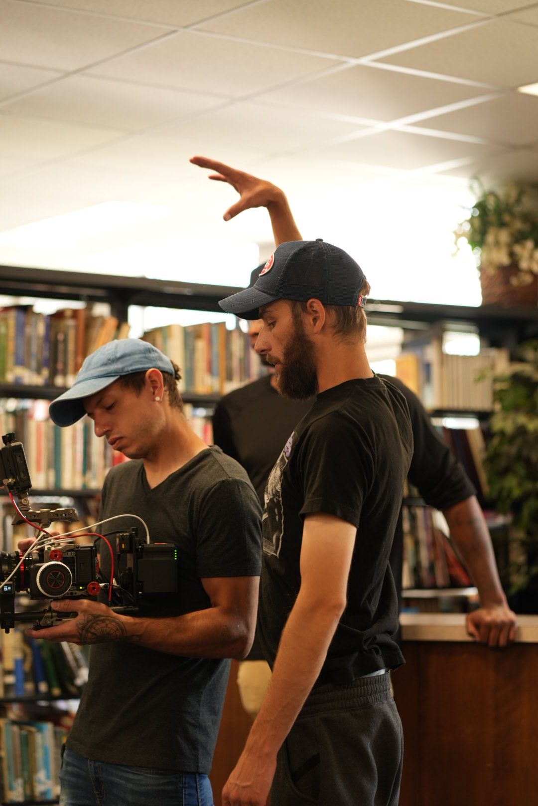 Two people in a library, one filming the other, who is gesturing with their hand, with bookshelves in the background.