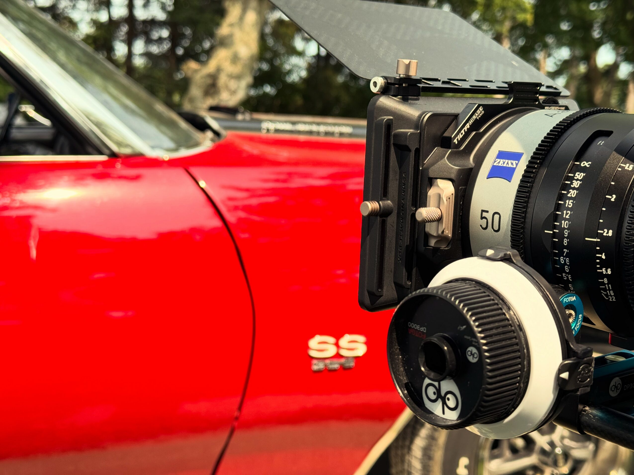 Close-up of a camera mounted on a tripod, capturing a red car with SS badge parked outdoors, trees in background.