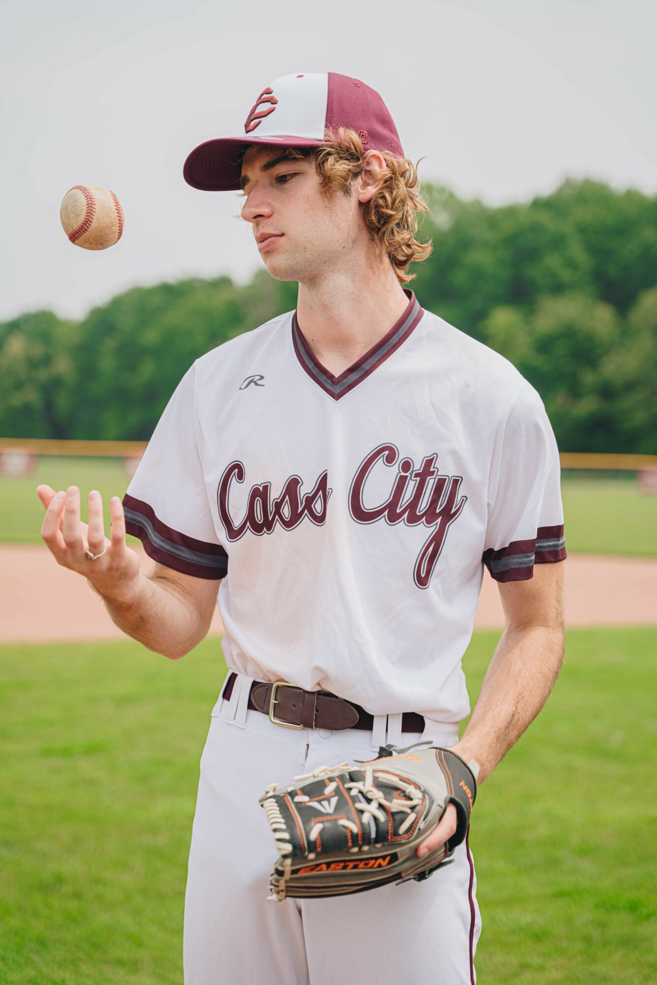 Young baseball player in uniform on field, holding glove, with baseball in air, trees in background.