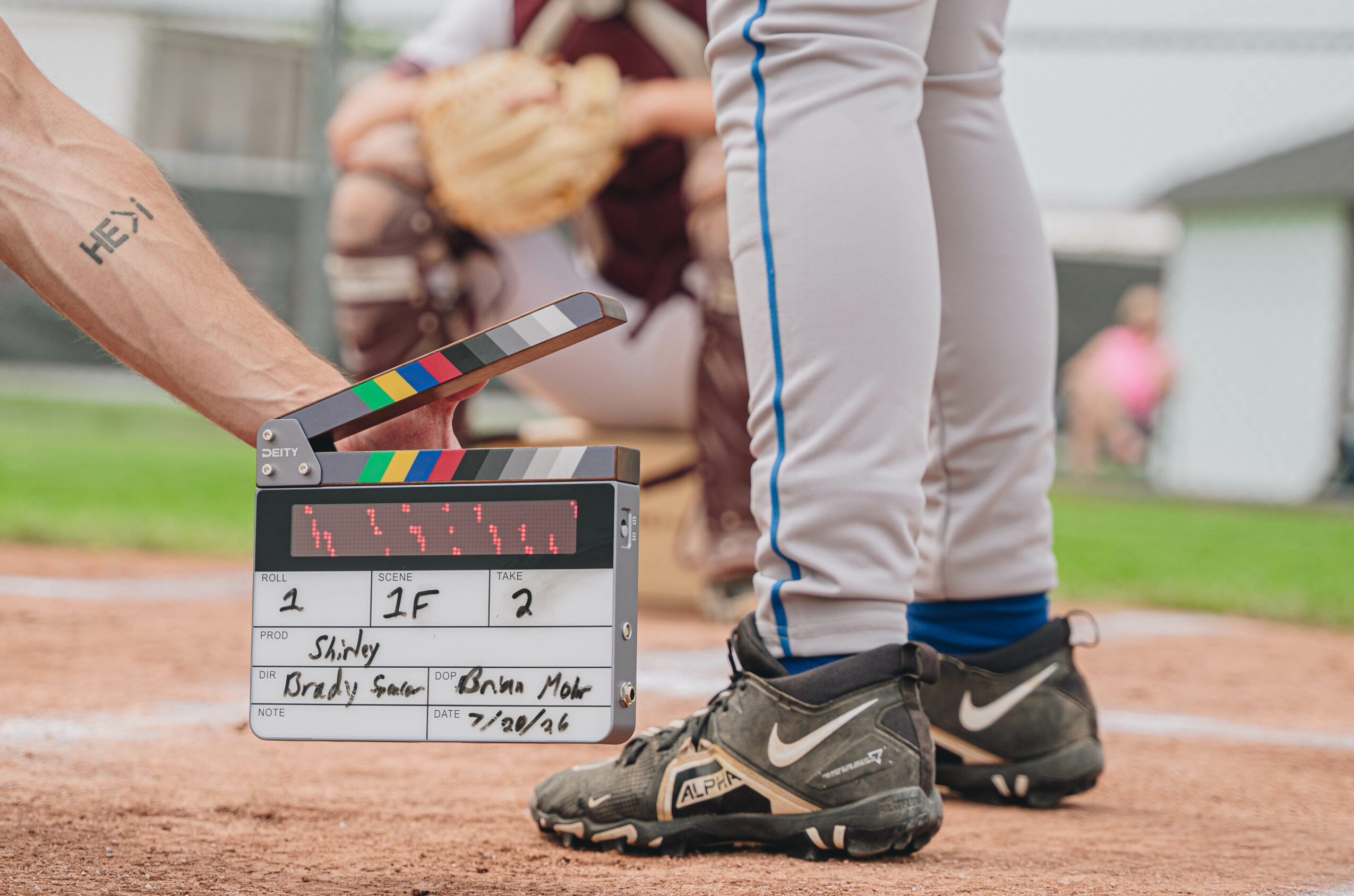 Close-up of a person wearing athletic shoes and gray pants on a sports field, with a clapperboard nearby, blurred background.