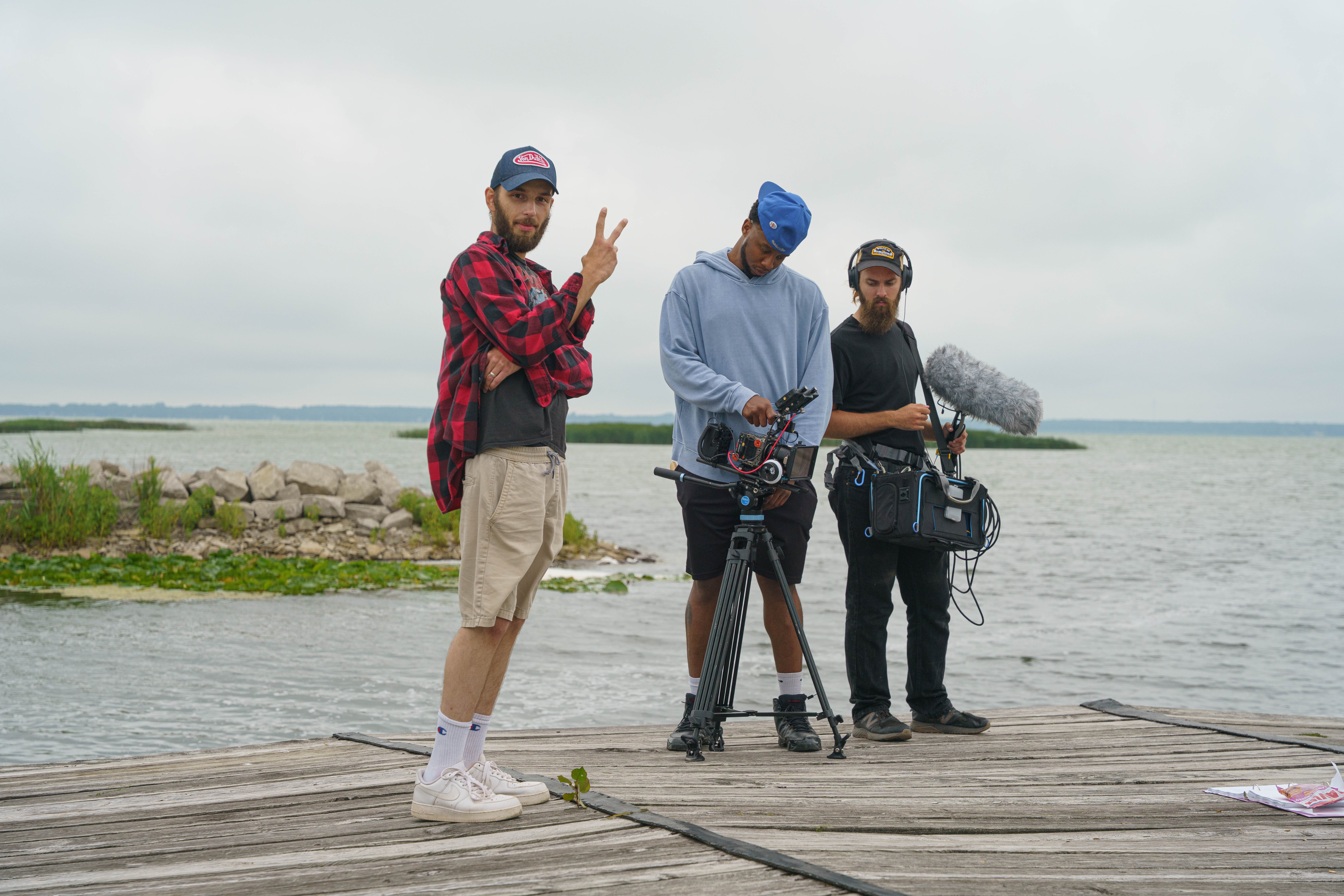 Three men stand on a wooden dock near water, one holding a microphone and recording equipment, others with cameras, overcast sky