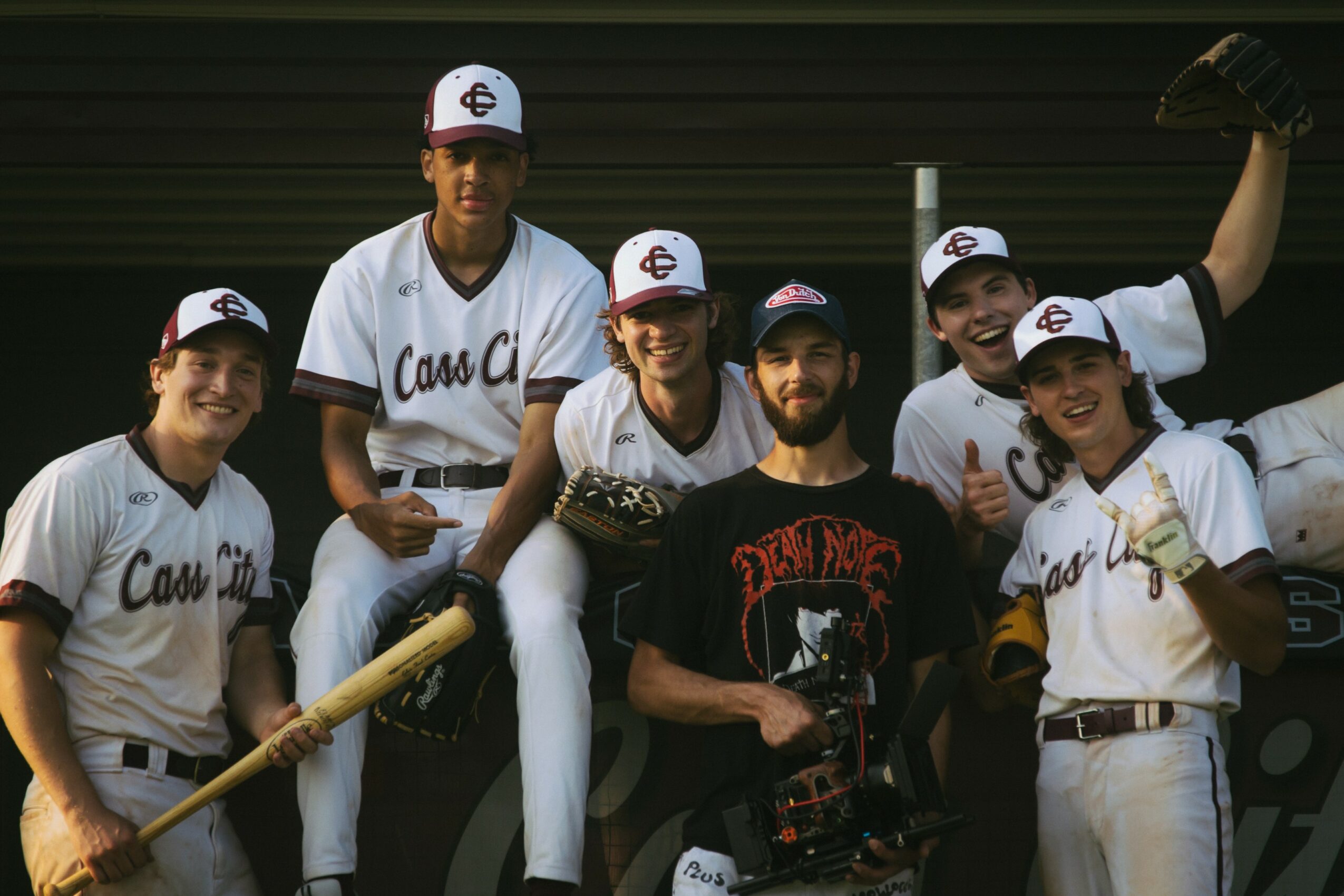 Group of six baseball players in uniforms and caps, smiling, posing outdoors against a dark background.