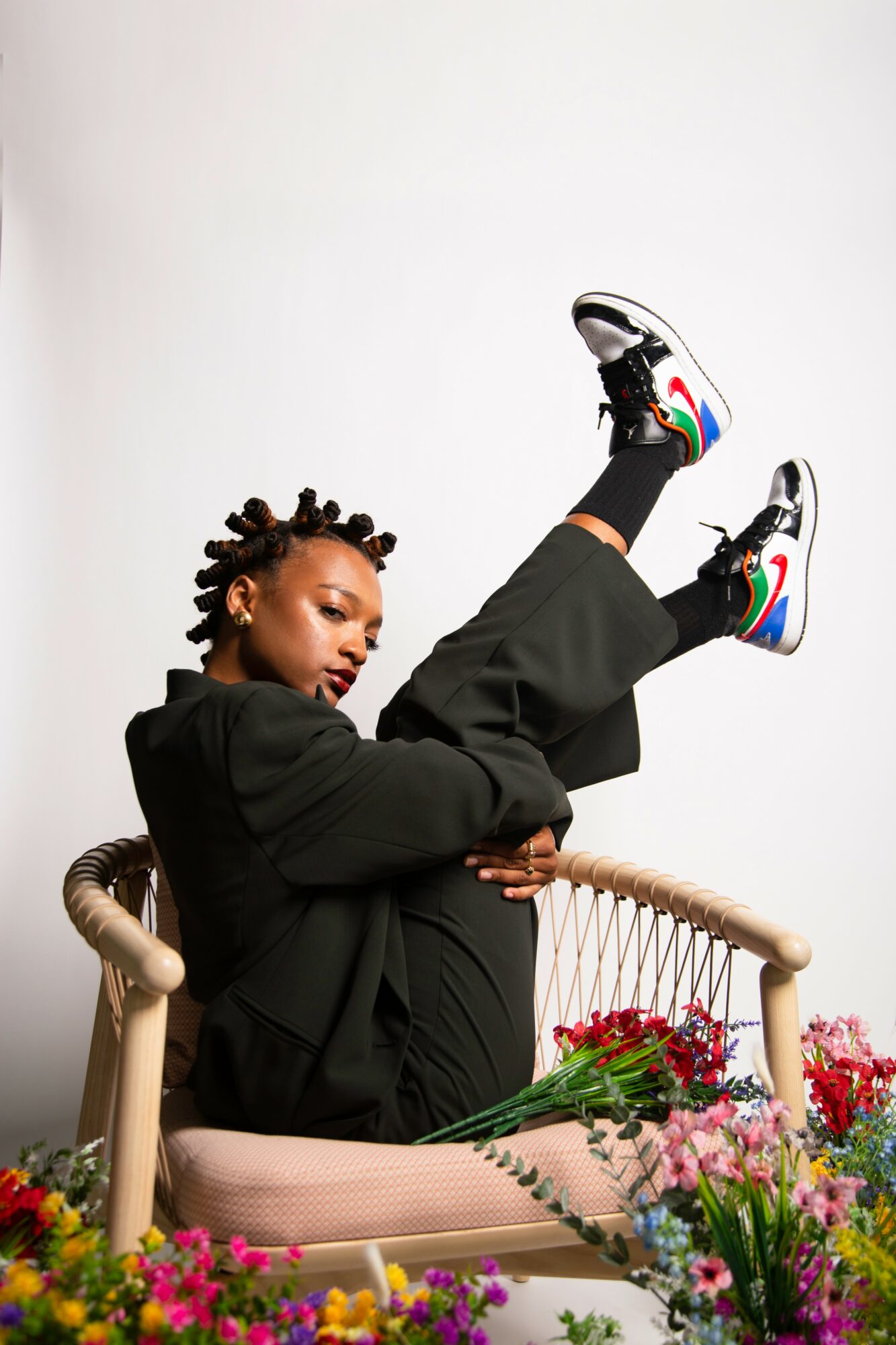 Person with dreadlocks in black clothing and colorful sneakers lying on a chair with flowers, upside down.