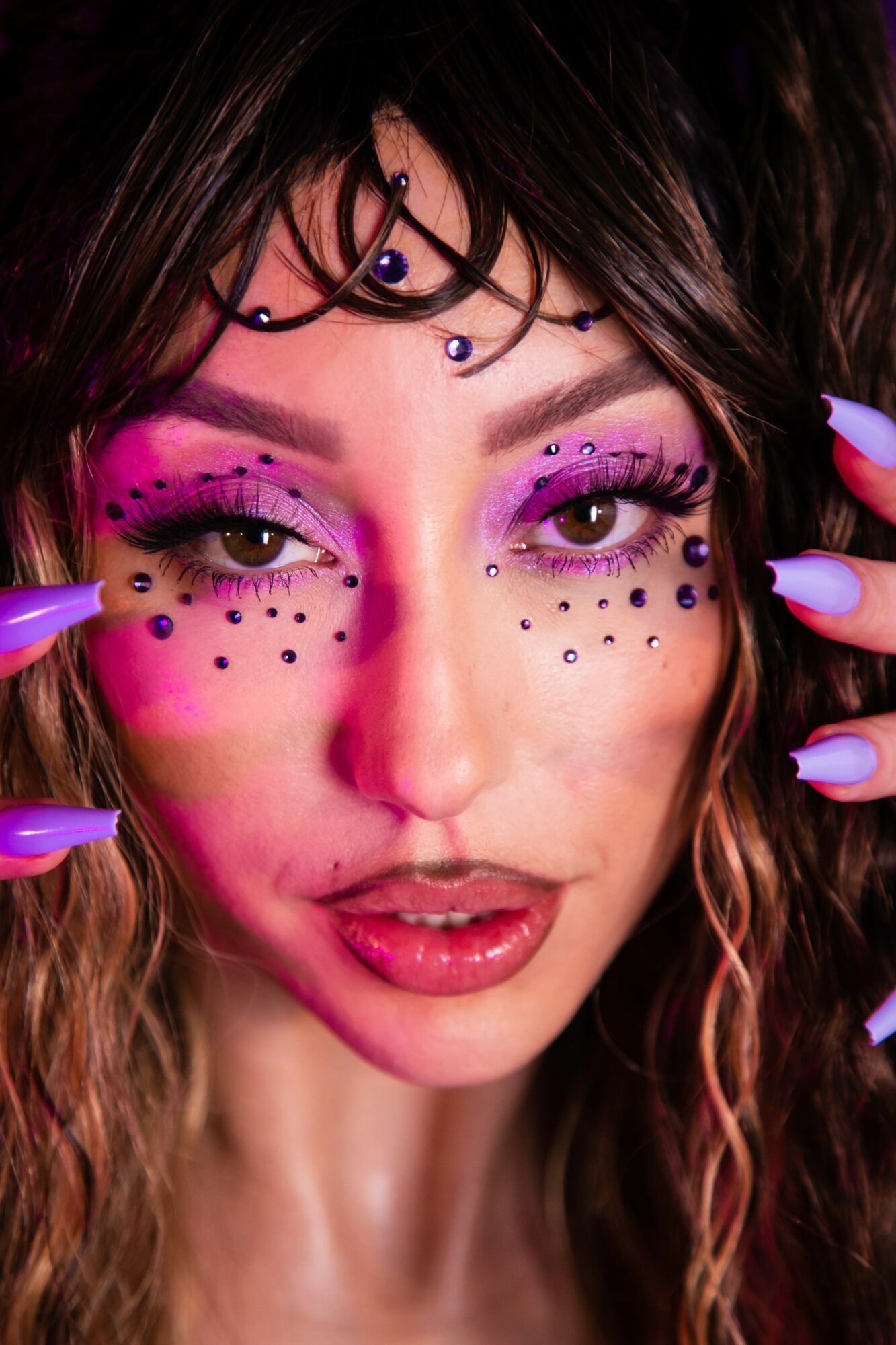 Close-up of a woman with colorful makeup, face decorations, and long nails, looking directly at the camera.