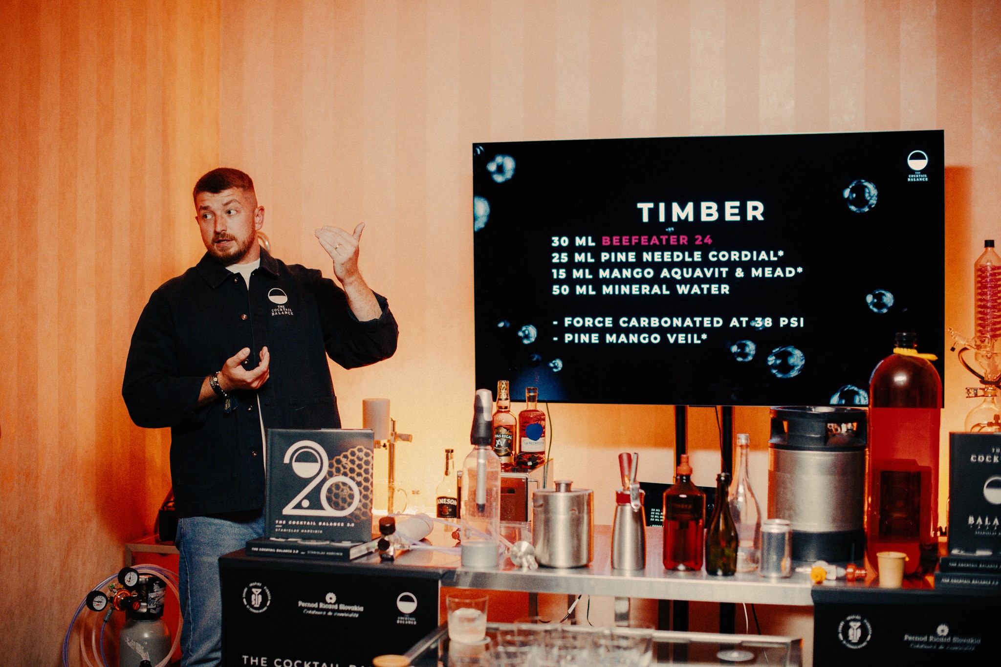 Person standing next to a table with drinks and a large screen displaying text, in a room with wooden walls.