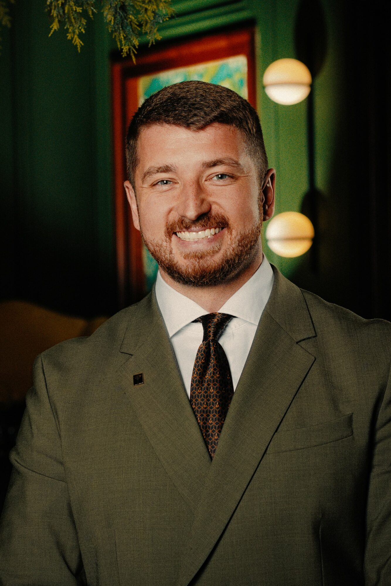 Man with short dark hair and beard smiling, wearing a suit and tie, in a decorated indoor setting.