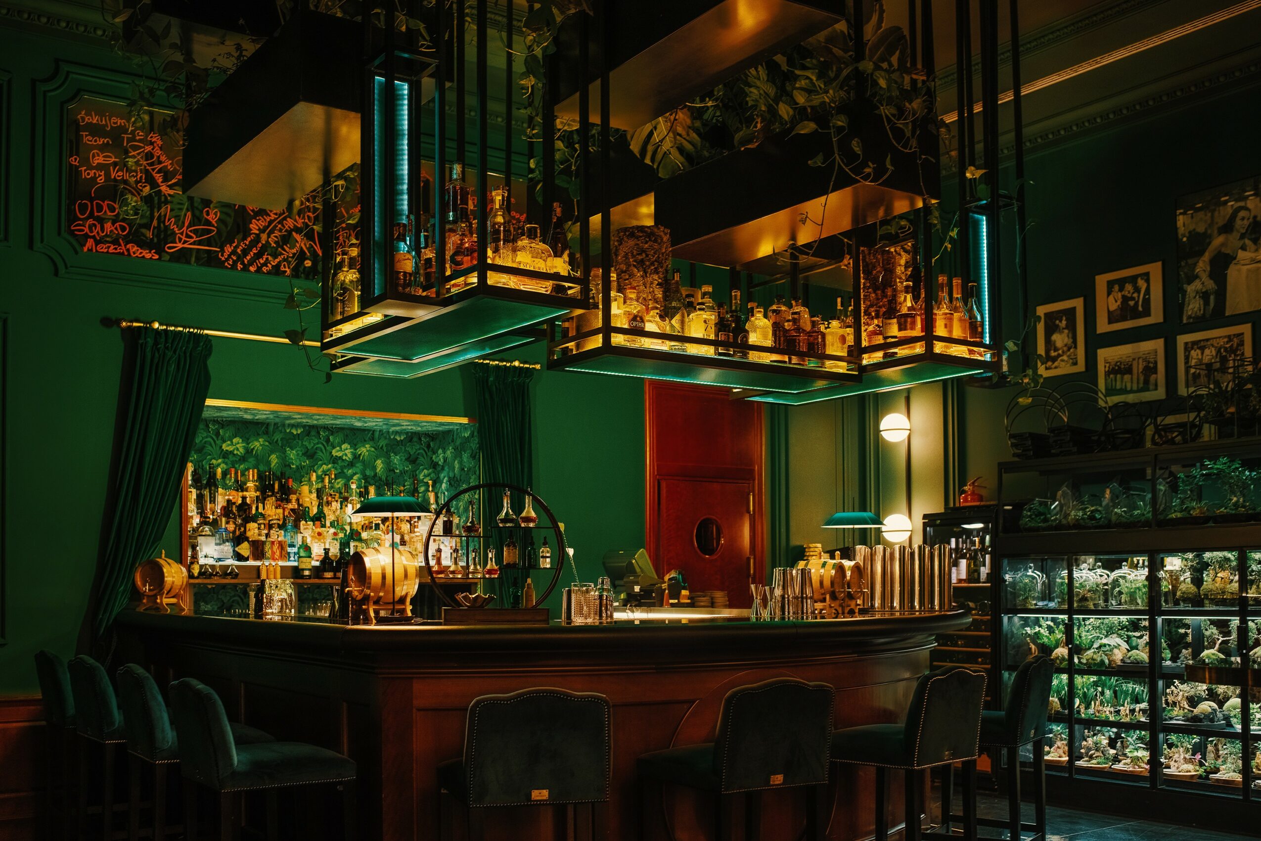 Bar with green walls, illuminated shelves, and hanging light fixtures, featuring bottles and plants, with seating area and display case.