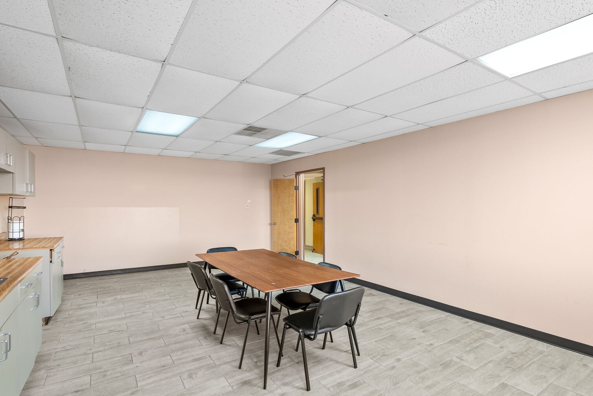 Empty room with a table and chairs, beige walls, ceiling tiles, and a door in the background.