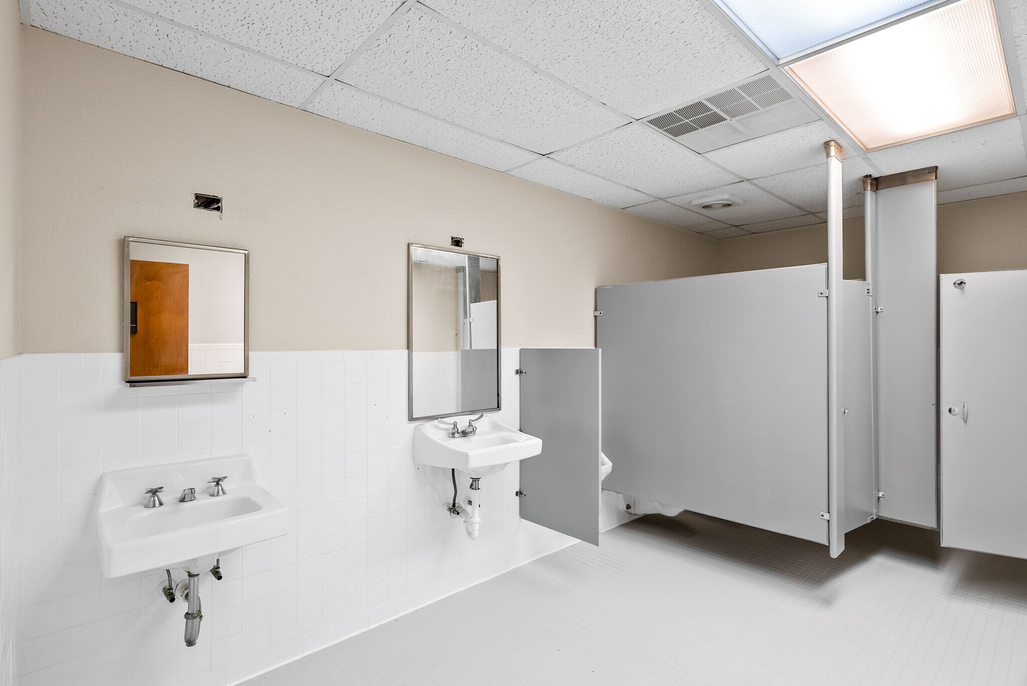 Public restroom with two sinks, mirrors, and partitioned stalls, white walls, ceiling tiles, and fluorescent lighting.