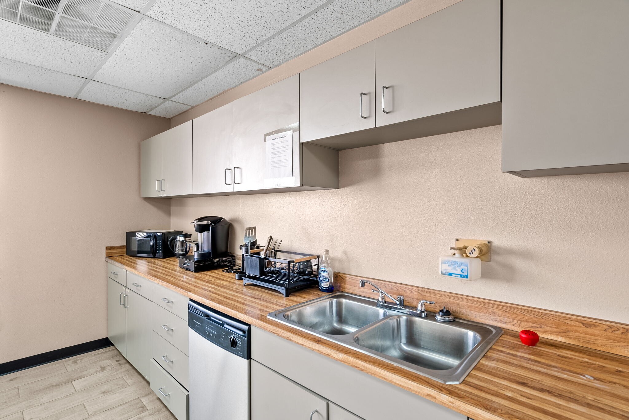 Kitchen counter with appliances, sink, and cabinets, beige wall, tiled ceiling, and wooden countertop.