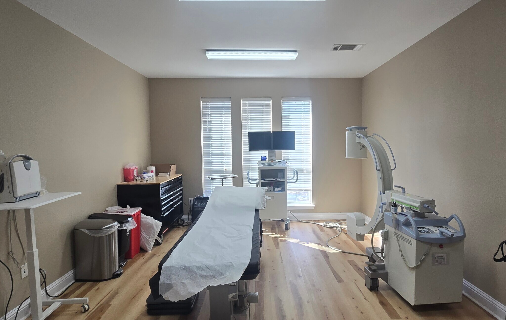Medical examination room with a bed, medical equipment, and windows with blinds, beige walls, and wooden floor.