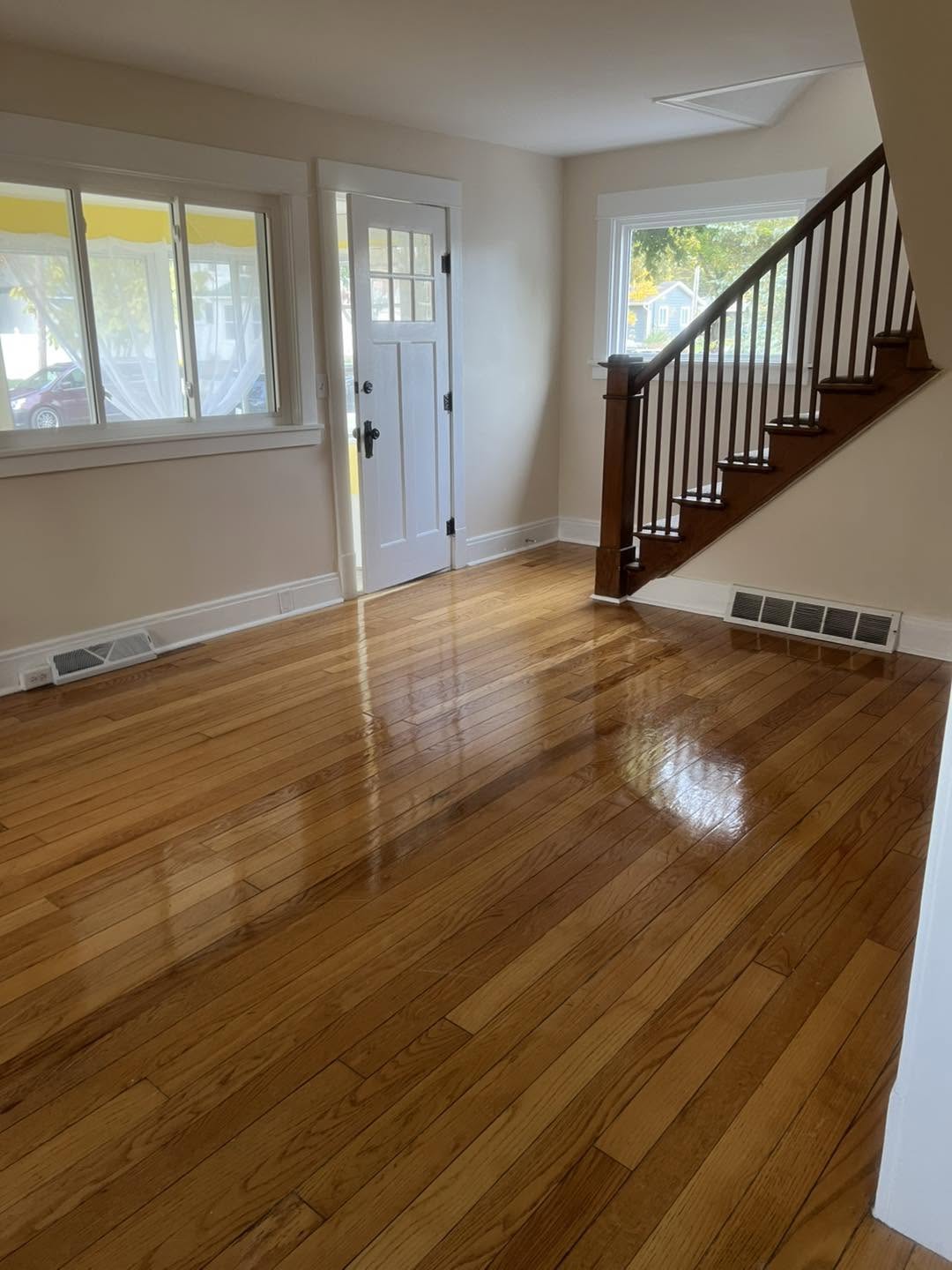 Empty living room with hardwood floor, front door, windows, and staircase with wooden railing, sunlight streaming in.