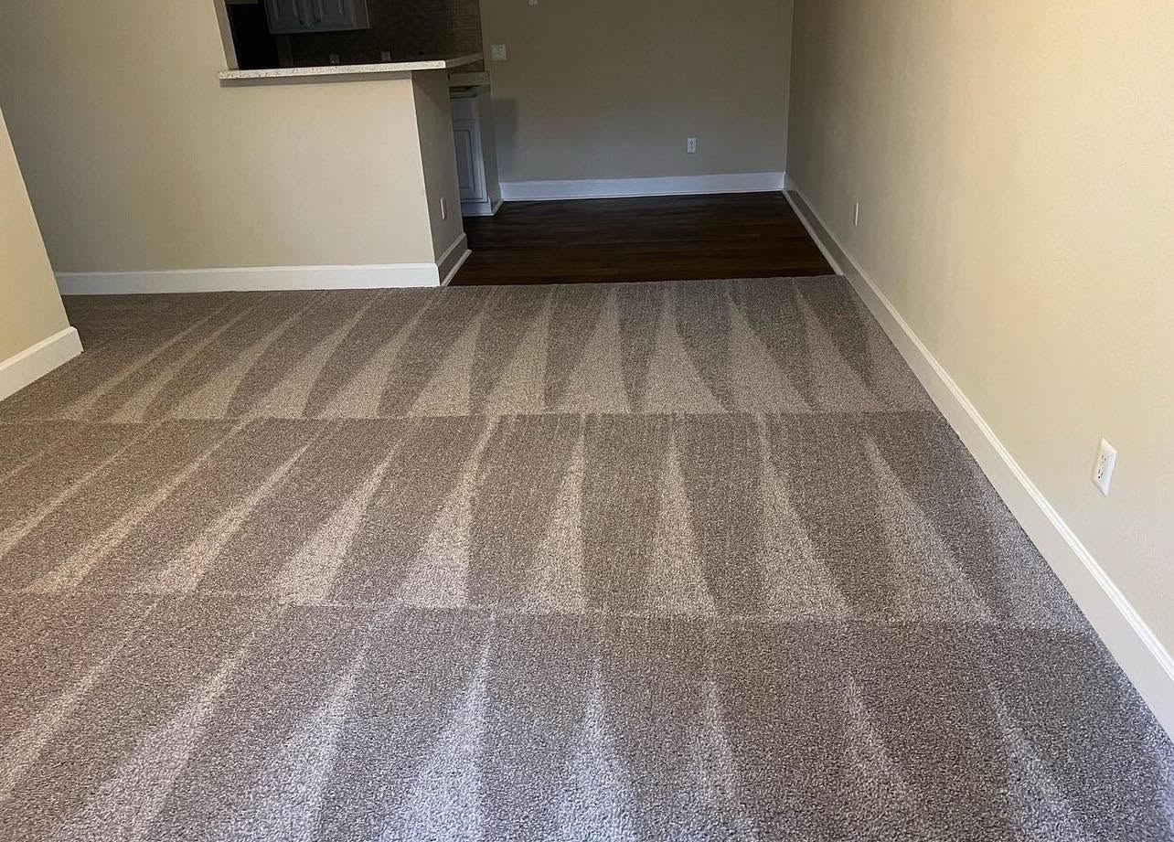 Carpeted room with striped pattern, beige walls, and a small section of dark hardwood floor in the background.