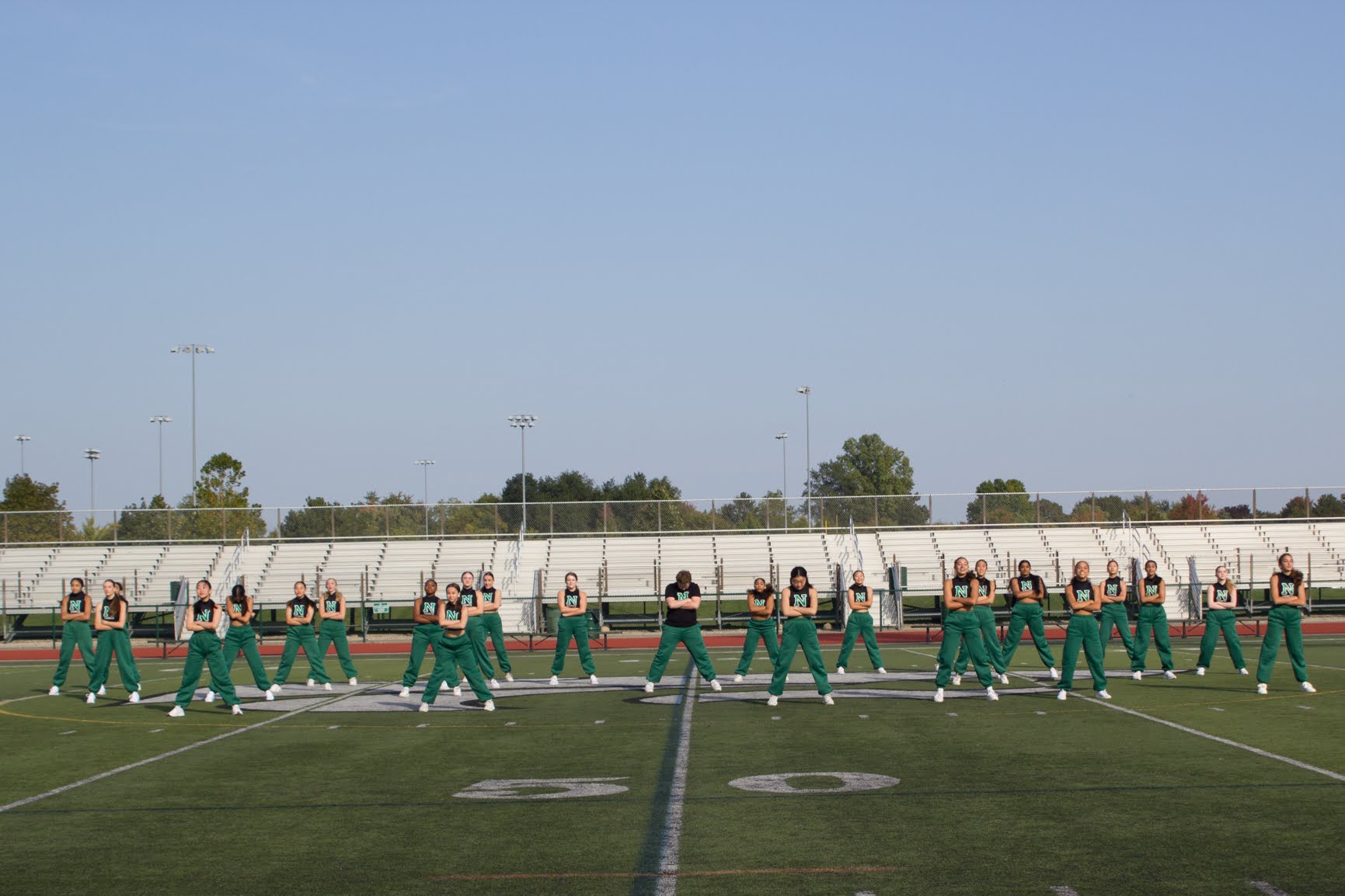 Group of cheerleaders performing a synchronized routine on a football field, with a clear sky and stadium seating in background.