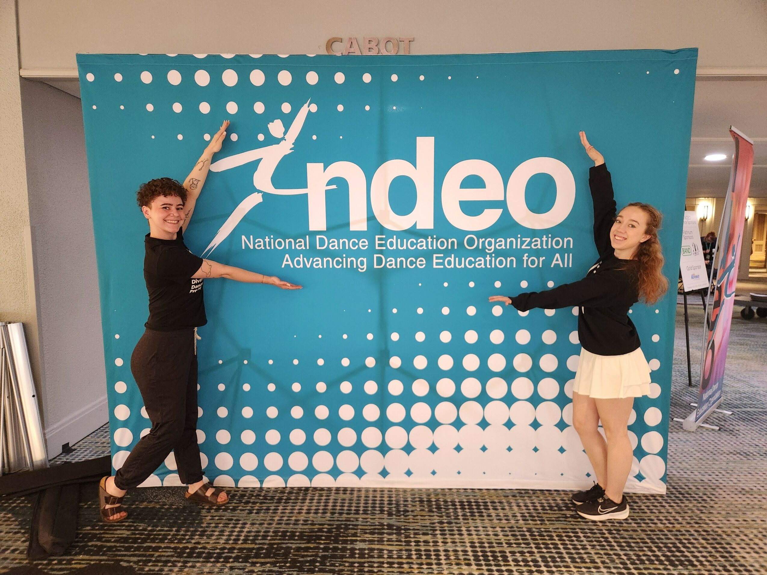 Two people standing in front of a blue backdrop with white dots and the text 'ndeo' and 'National Dance Education Organization'.