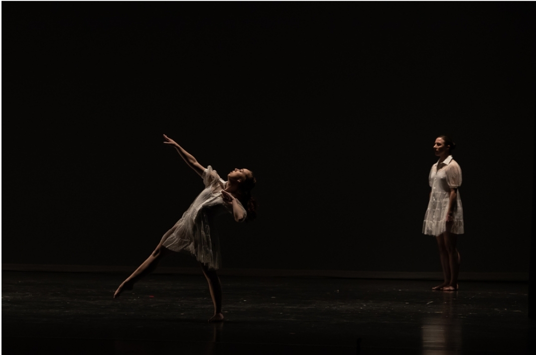 Two dancers perform on a dark stage, one in a pose with arms extended and the other standing in the background.