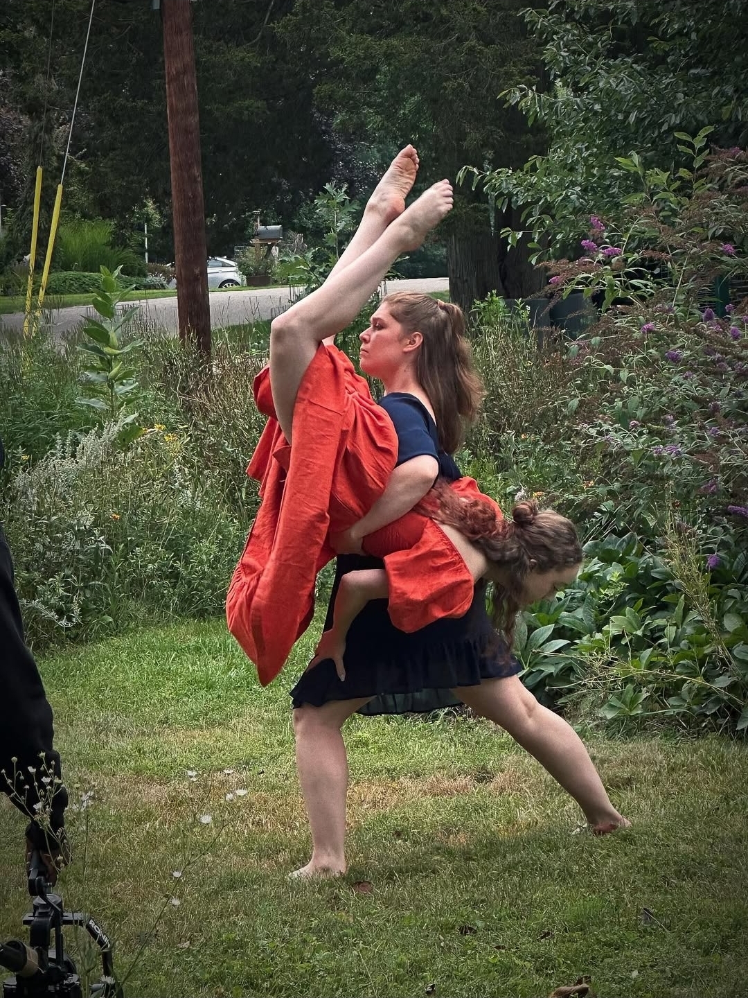 Two children playing outdoors, one standing and the other leaning on her back, with trees and plants in the background.