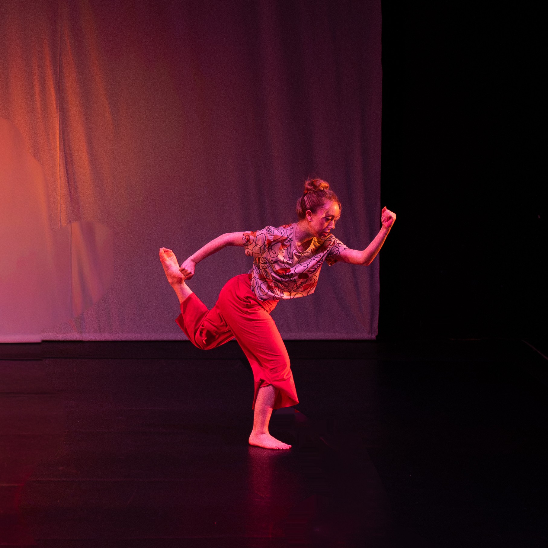 Person performing a dance move on stage with a colorful background, wearing a patterned top and red pants.