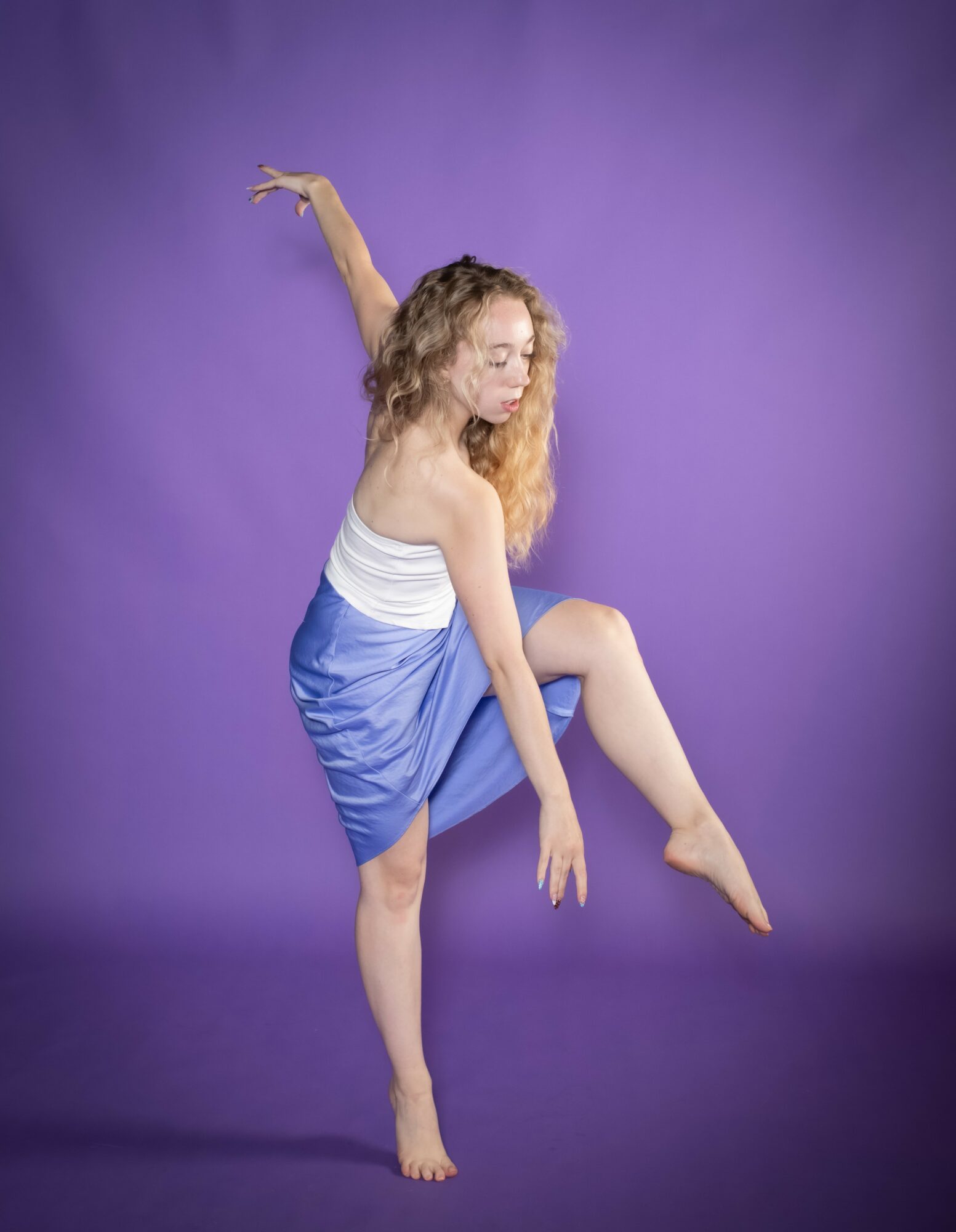 A woman with curly hair dances against a purple background, wearing a white top and blue skirt, with one arm raised.