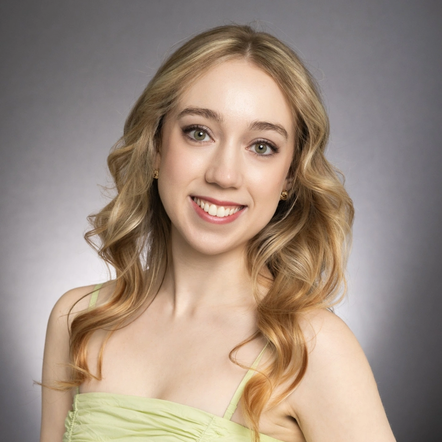 Young woman with wavy blonde hair smiling, wearing a light green strapless top, against a gray background.