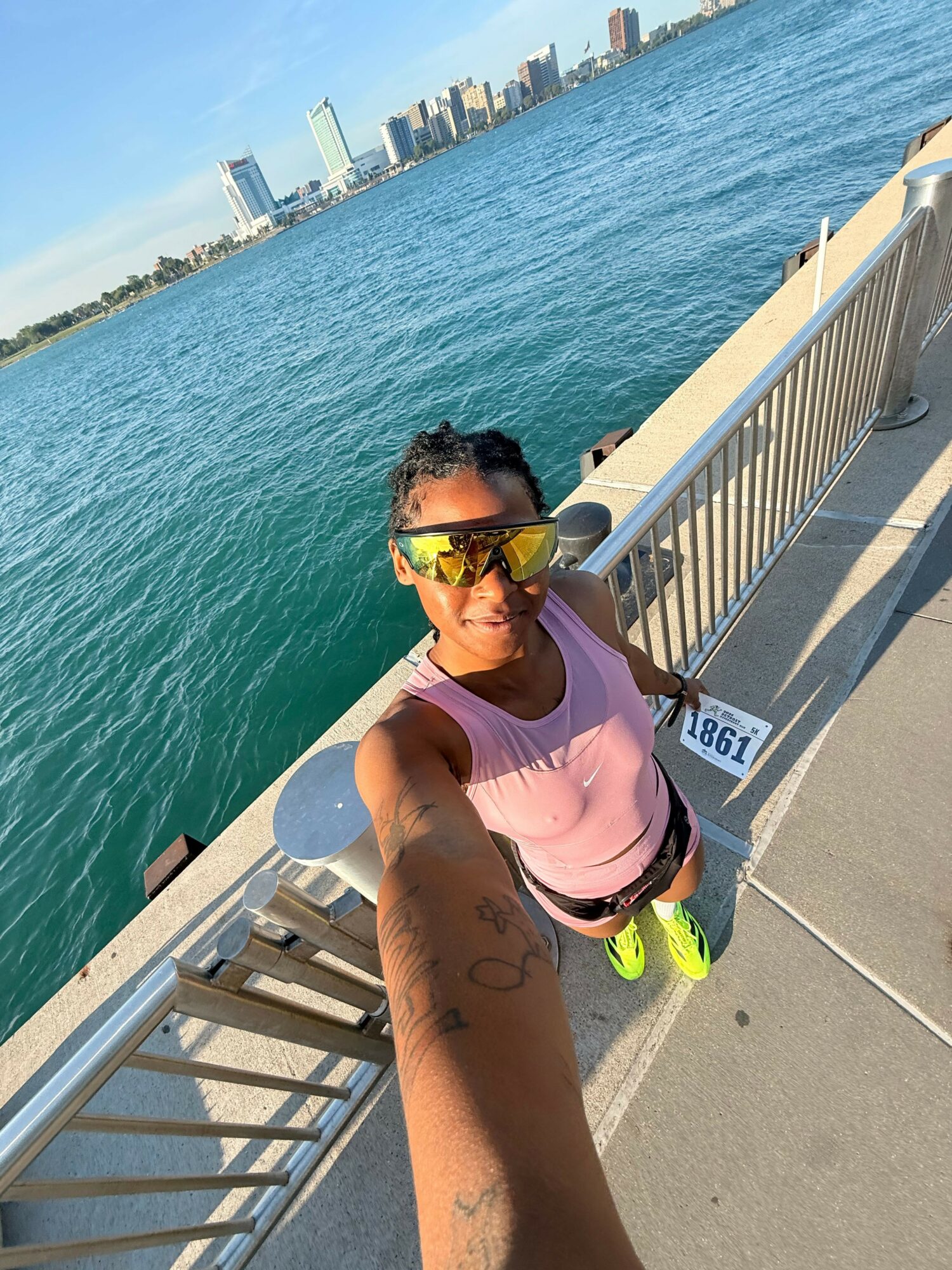 Person lying on a dock near water, wearing sunglasses and a pink shirt, with a tattooed arm extended towards the camera.