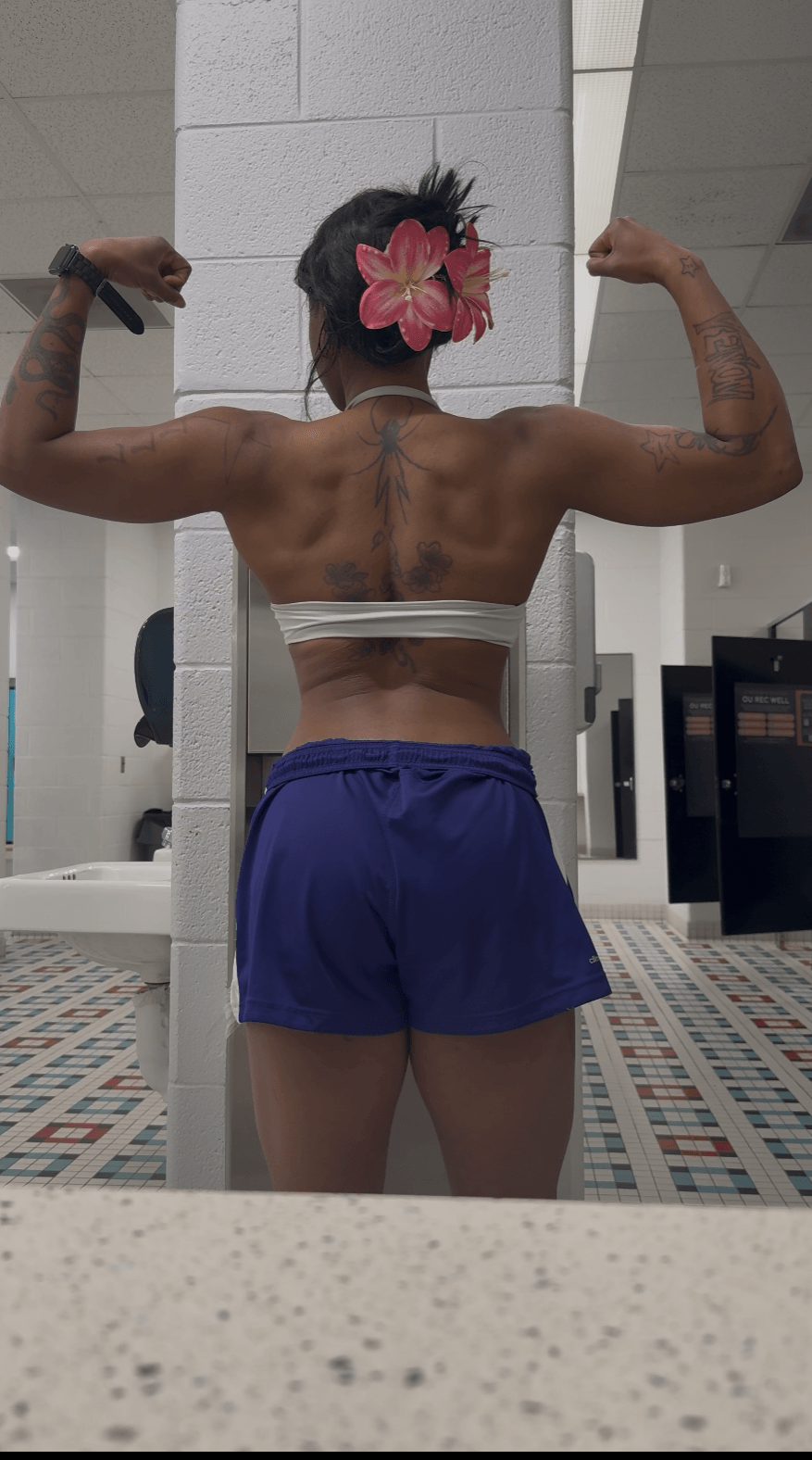 Person with a flower in hair flexing back muscles in a gym, facing mirror, wearing white top and blue shorts.