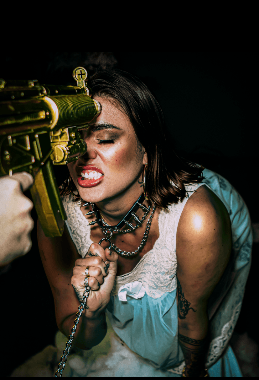 Woman with dark hair and makeup aiming a yellow gun, wearing jewelry and a blue and white outfit, against a dark background.