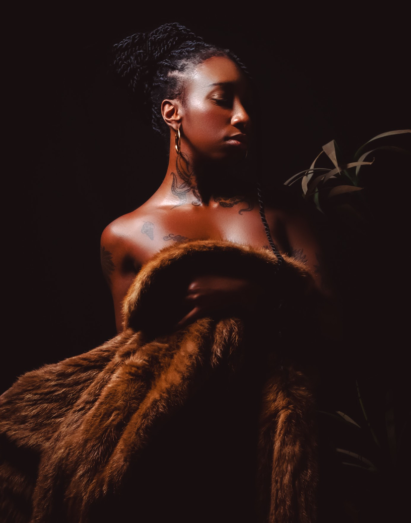 A woman with braided hair and earrings, draped in a fur coat, poses against a dark background.