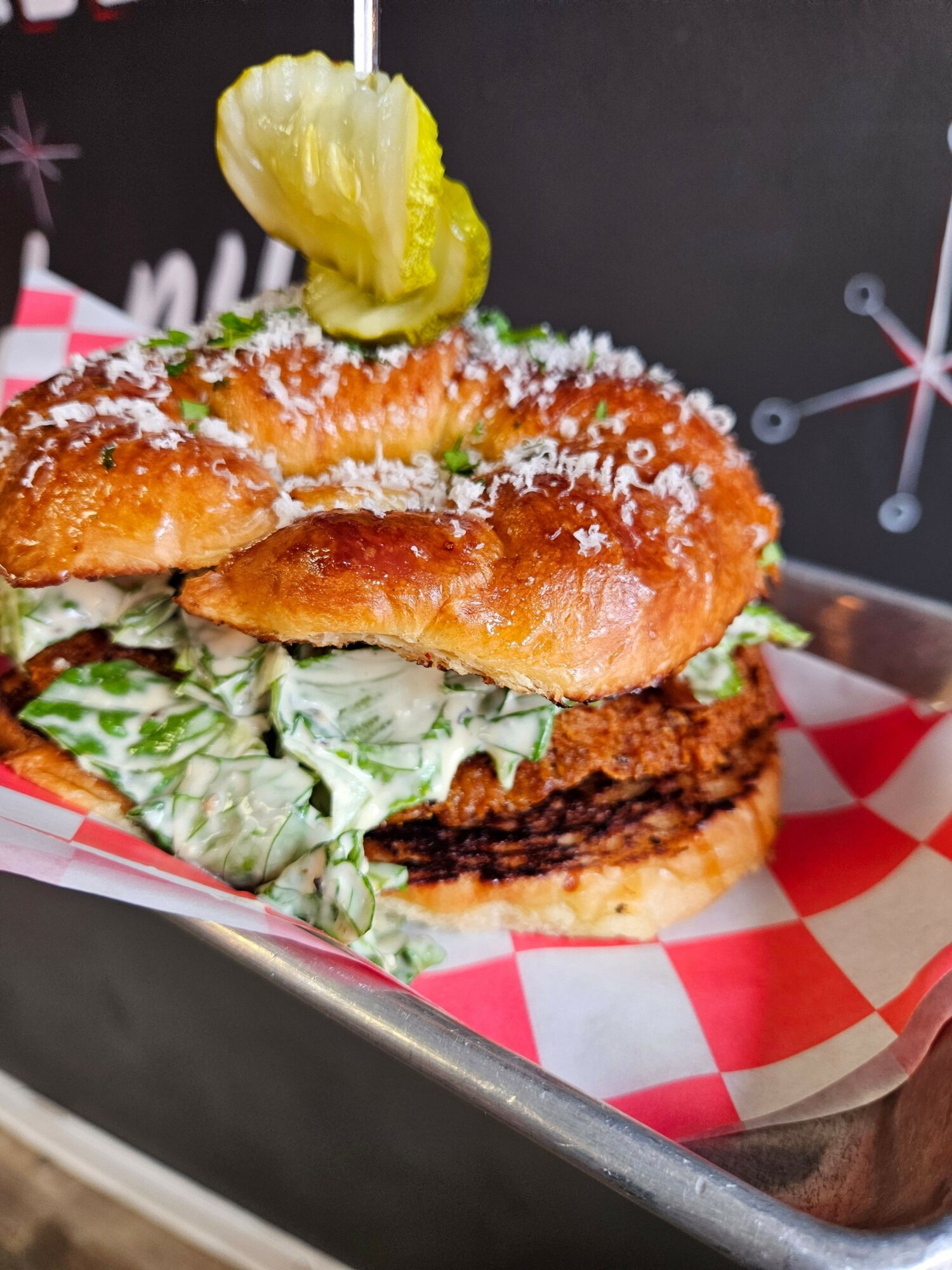 Fried fish sandwich with lettuce, pickles, and a bun, served in a paper-lined basket with red and white checkered paper.