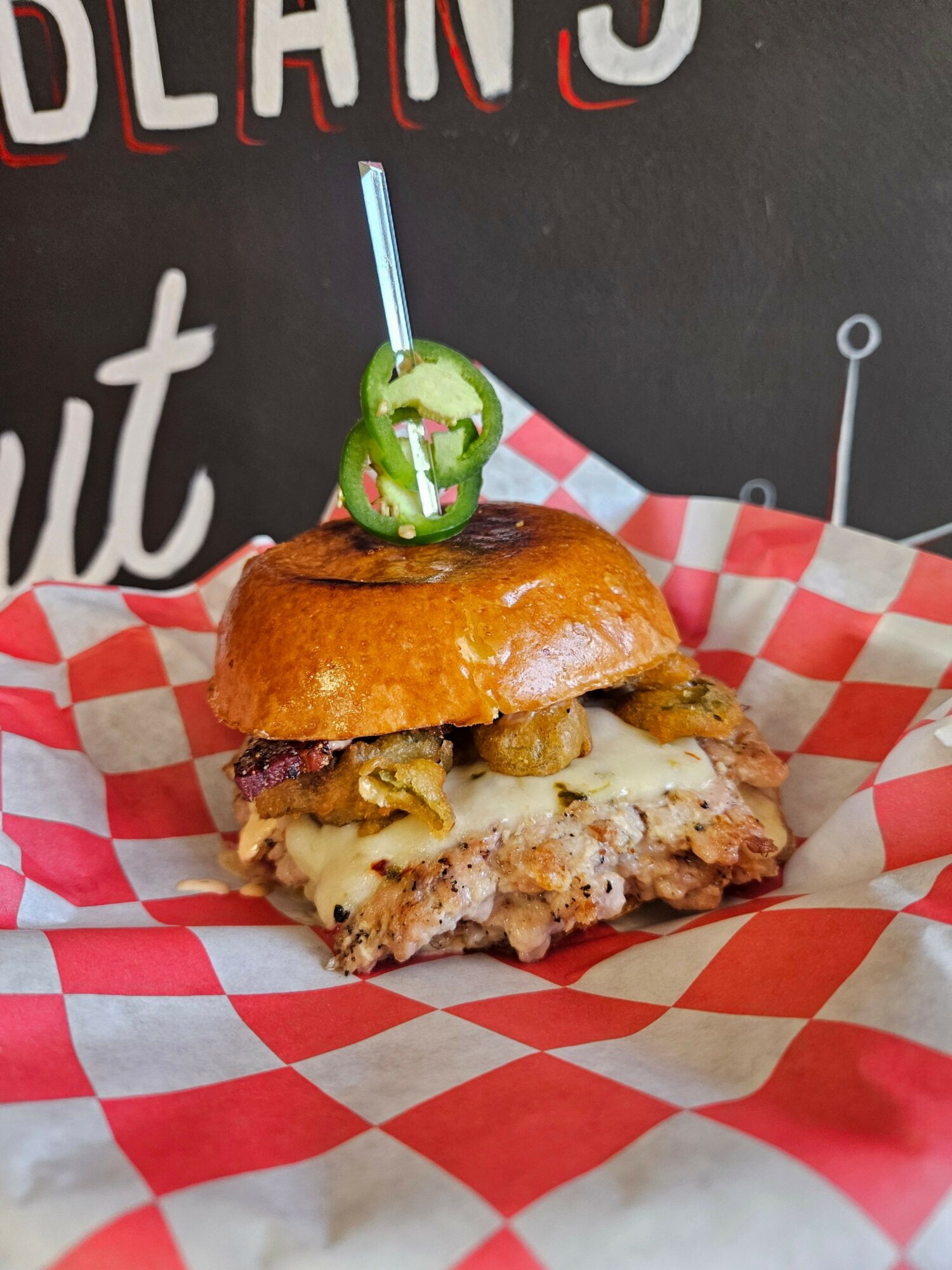 Sandwich with fried chicken, cheese, and toppings on red and white checkered paper, on a black surface with white text.